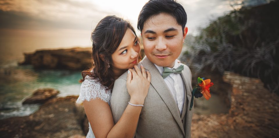 A woman in a white dress stands close to a man in a beige suit with a flower boutonniere, against a coastal background.