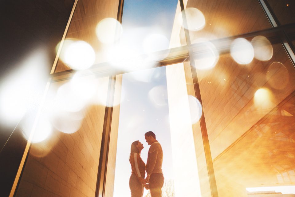 A couple stands holding hands, silhouetted against a modern glass building with warm sunlight and soft bokeh lights in the foreground.