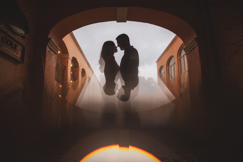 Silhouette of a couple facing each other under an archway with a bright background, captured through a reflective surface creating a symmetrical effect.