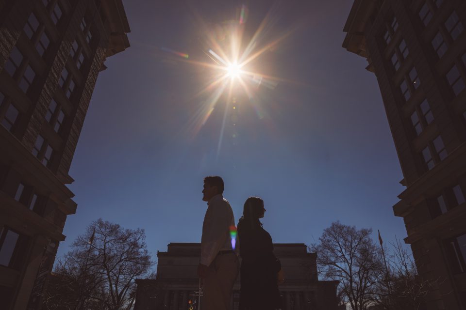 Two people stand back-to-back between two tall buildings. The sun shines brightly at the top center of the image.