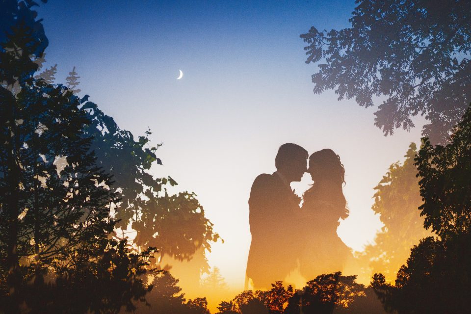 Silhouetted couple kissing against a sunset backdrop, surrounded by trees and a crescent moon in the sky.