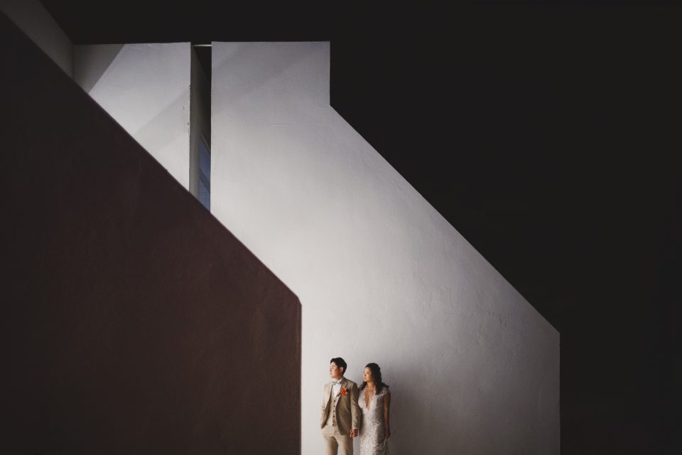 A couple in formal attire stands against a modern, angular white building at night, with dramatic lighting highlighting their presence.