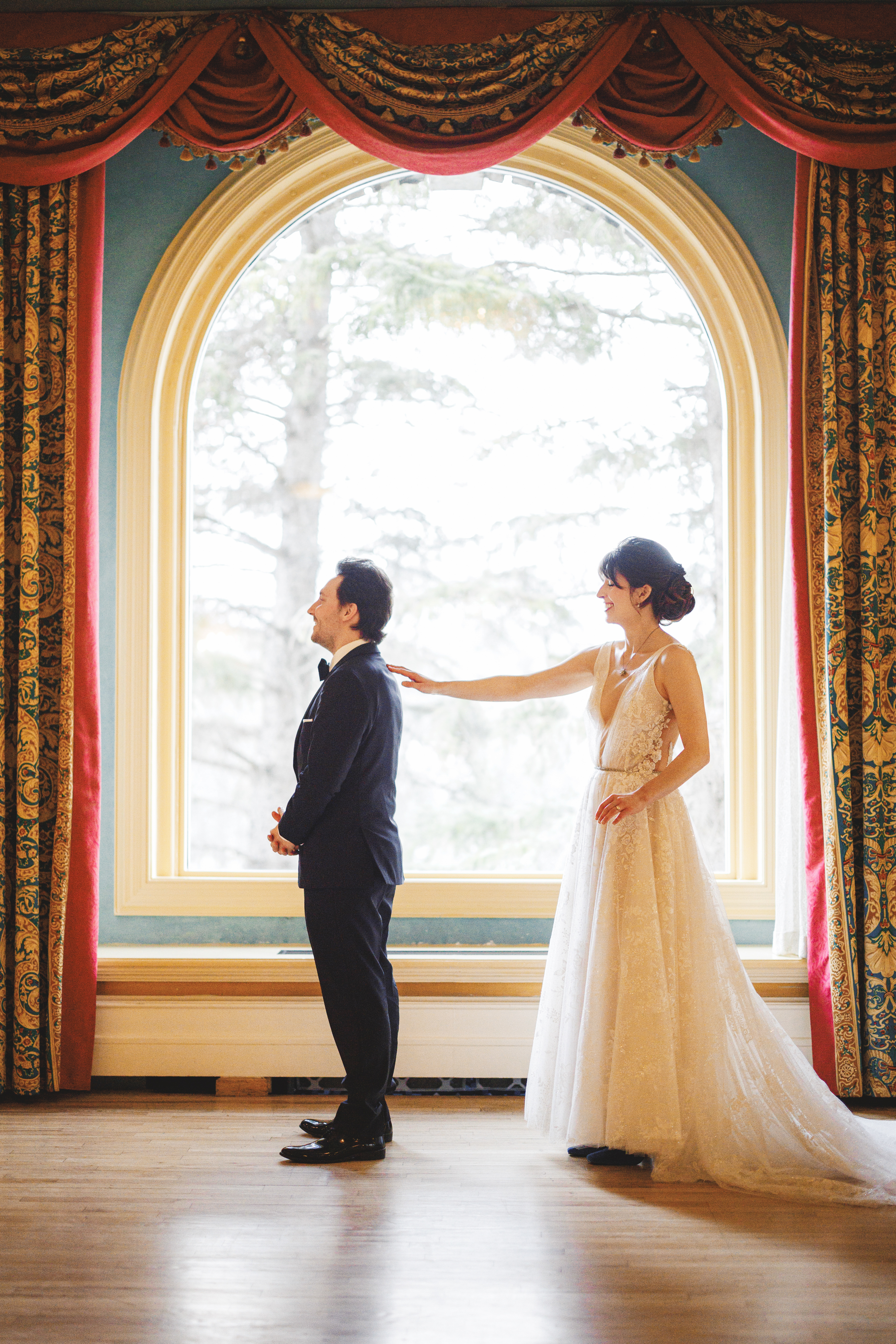 A bride in a white dress stands behind a groom in a suit, gently touching his shoulder in front of a large arched window with ornate curtains.