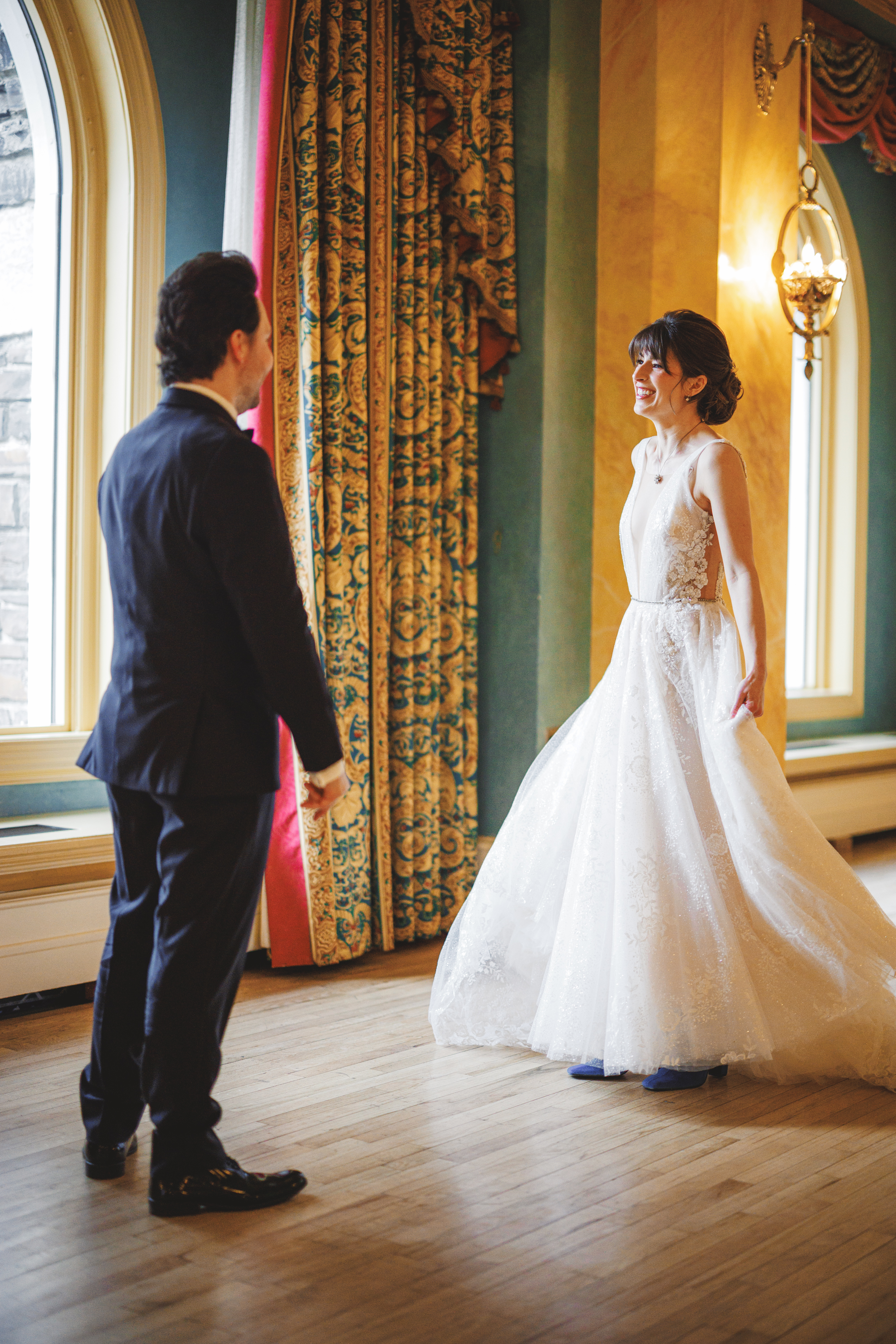 A bride in a white dress and a groom in a dark suit stand facing each other in an elegant room with large windows and ornate curtains.