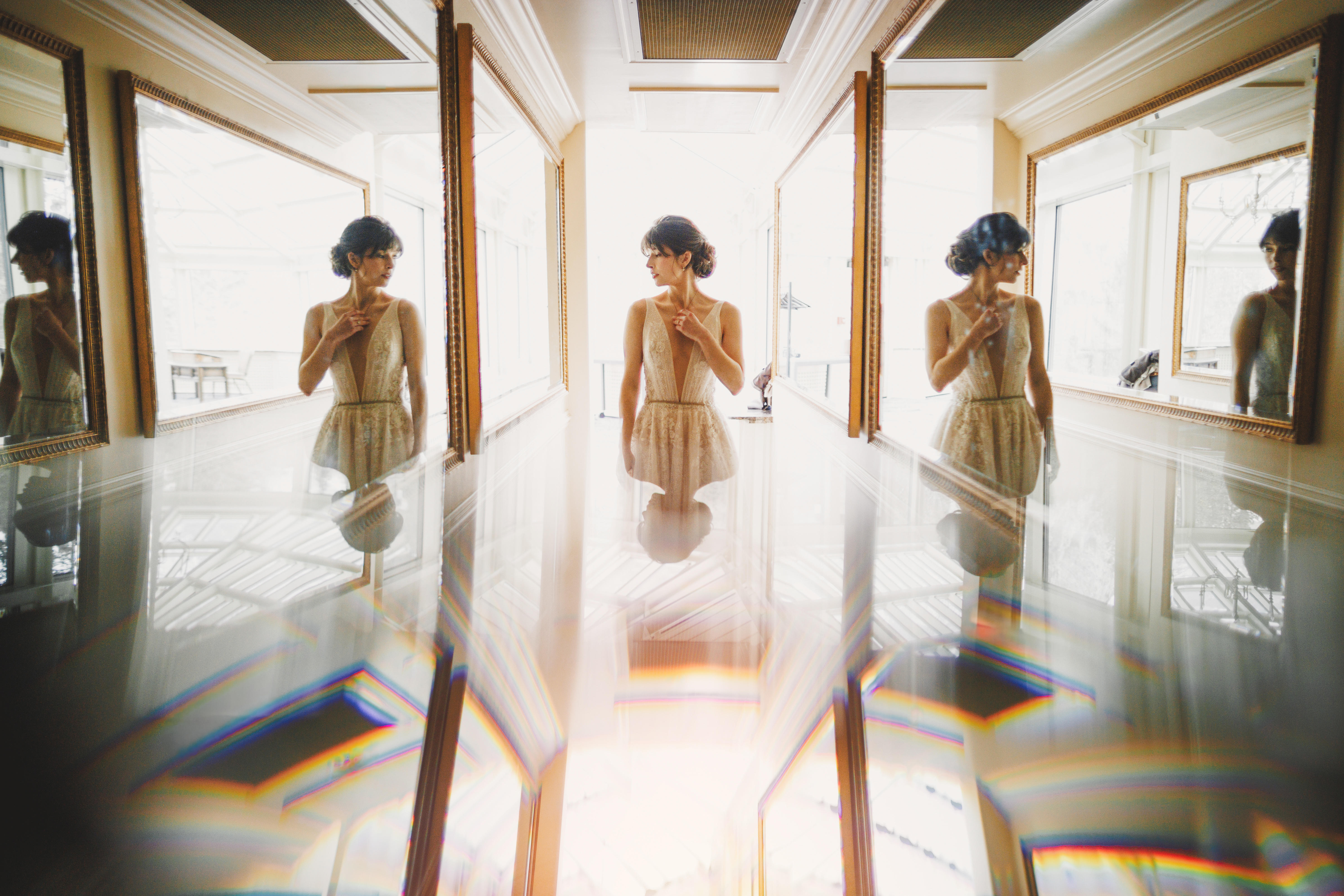 A woman in a white dress stands indoors between mirrors, with reflections and prism light effects visible on the glossy surface in front of her.