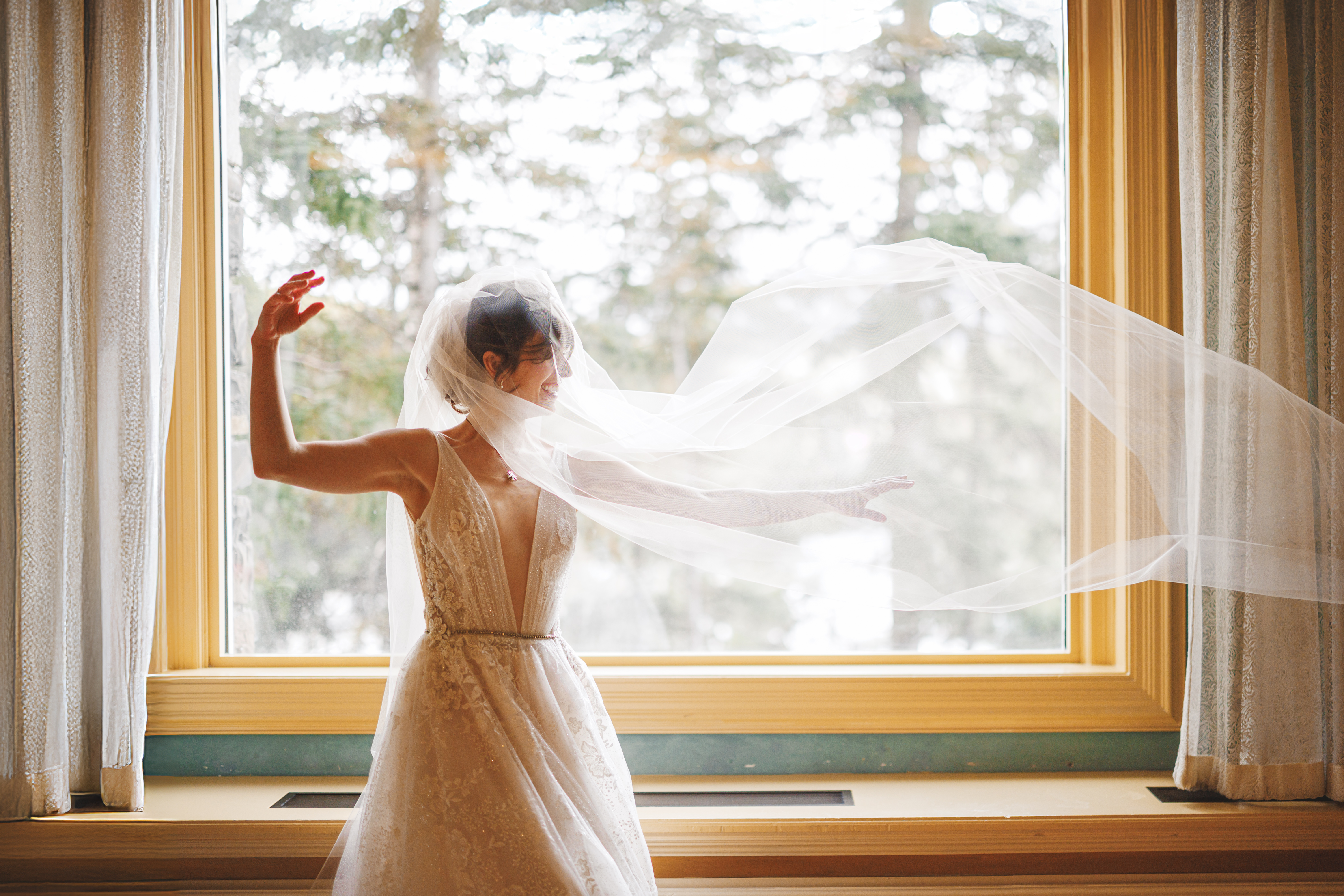 A bride in a white wedding dress stands in front of a large window, her veil flowing in the air as she raises one arm.
