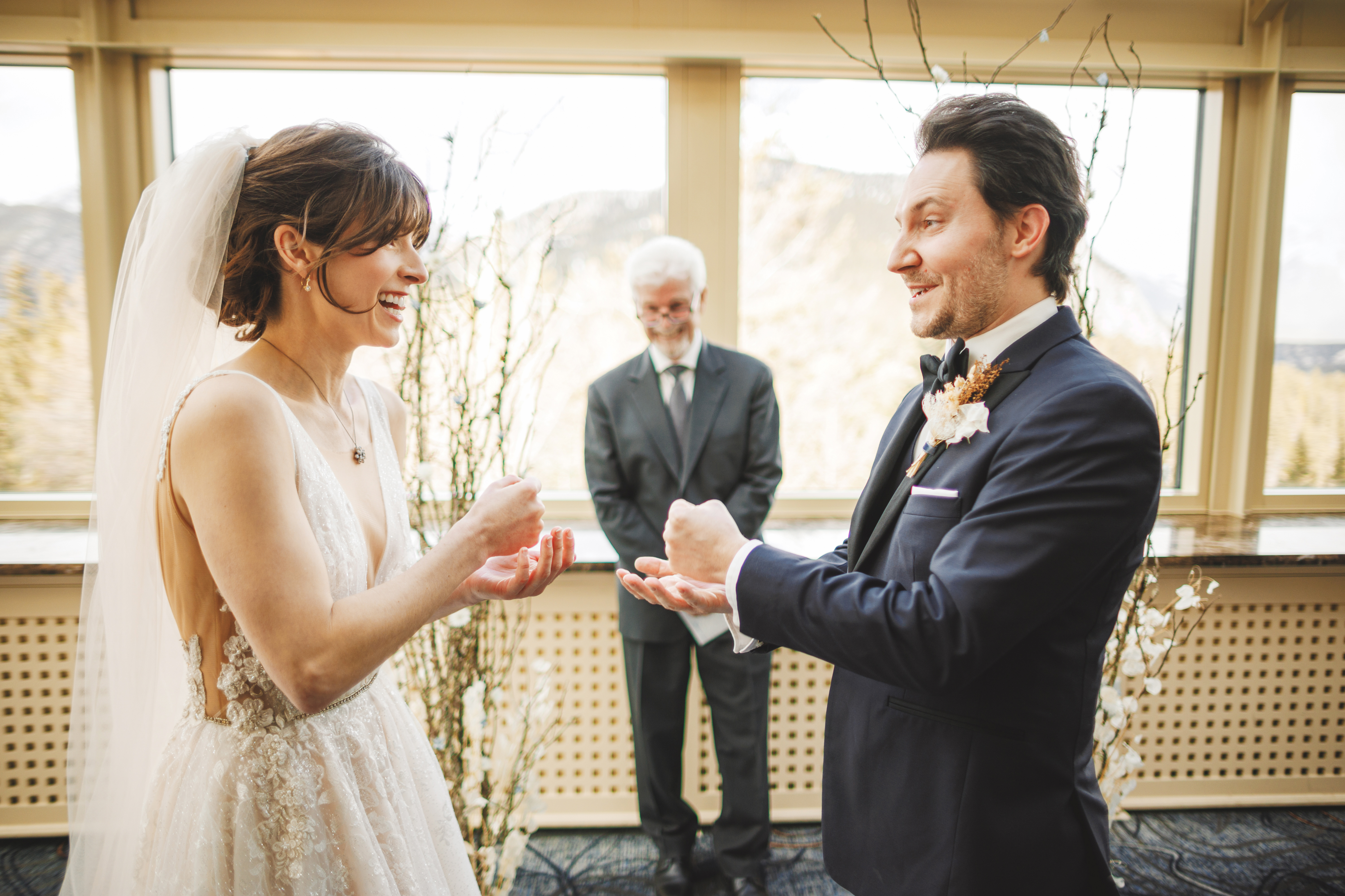 A bride and groom in wedding attire play a game of rock-paper-scissors, smiling, with an officiant standing behind them indoors.