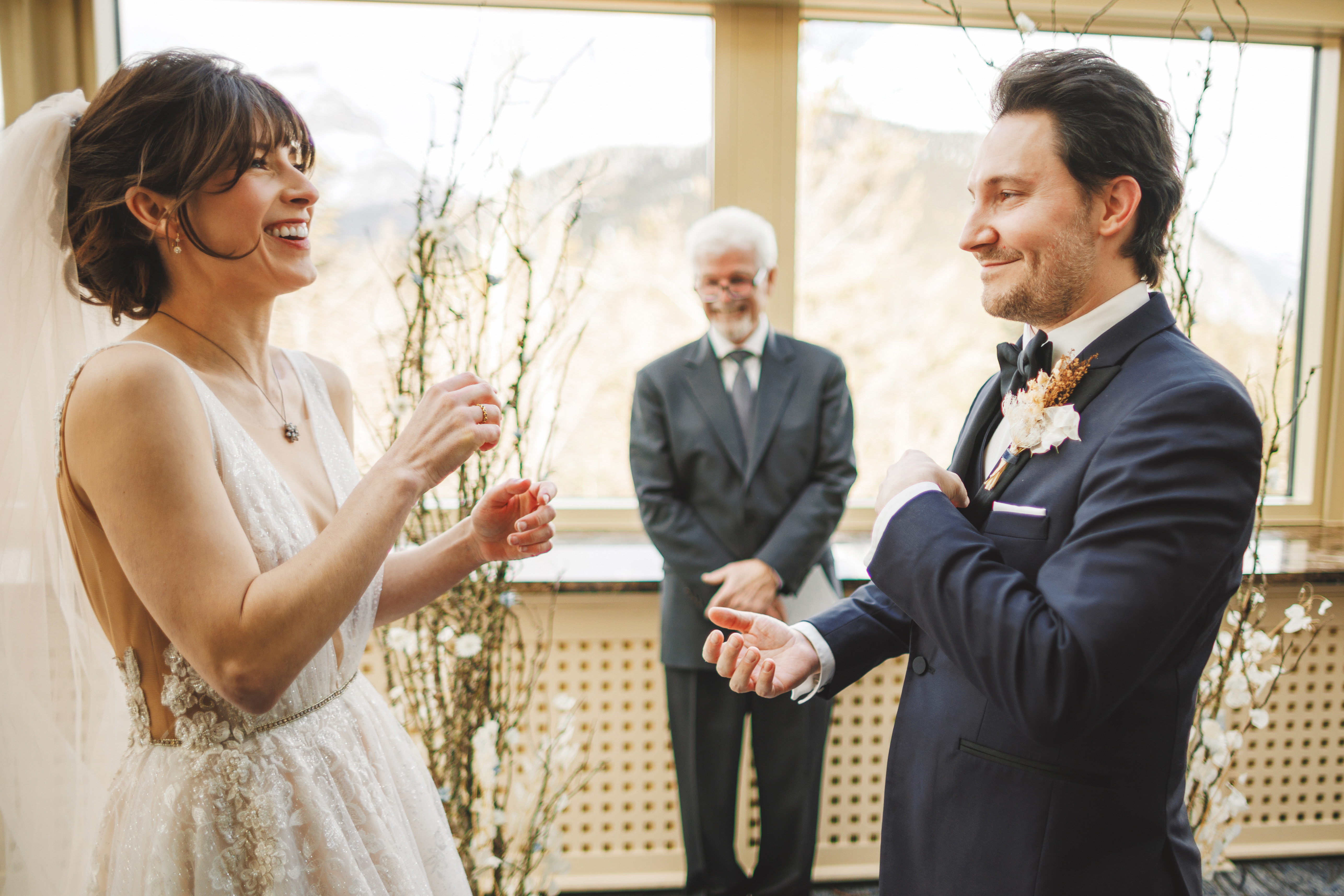 A bride and groom laugh during their wedding ceremony as an officiant stands behind them in a bright, window-filled room.