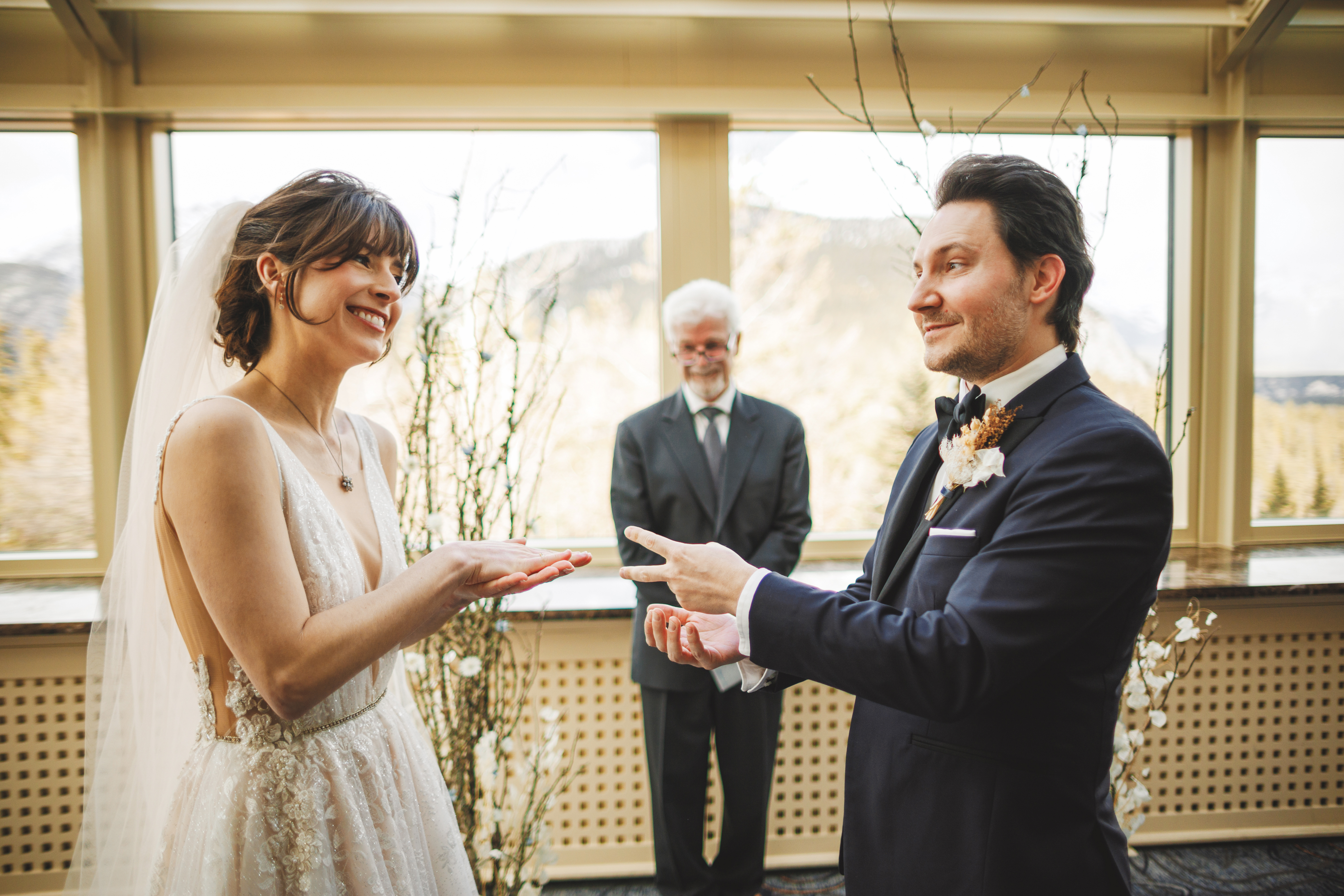 A bride and groom play rock-paper-scissors during their wedding ceremony, whilst an officiant stands behind them, smiling.