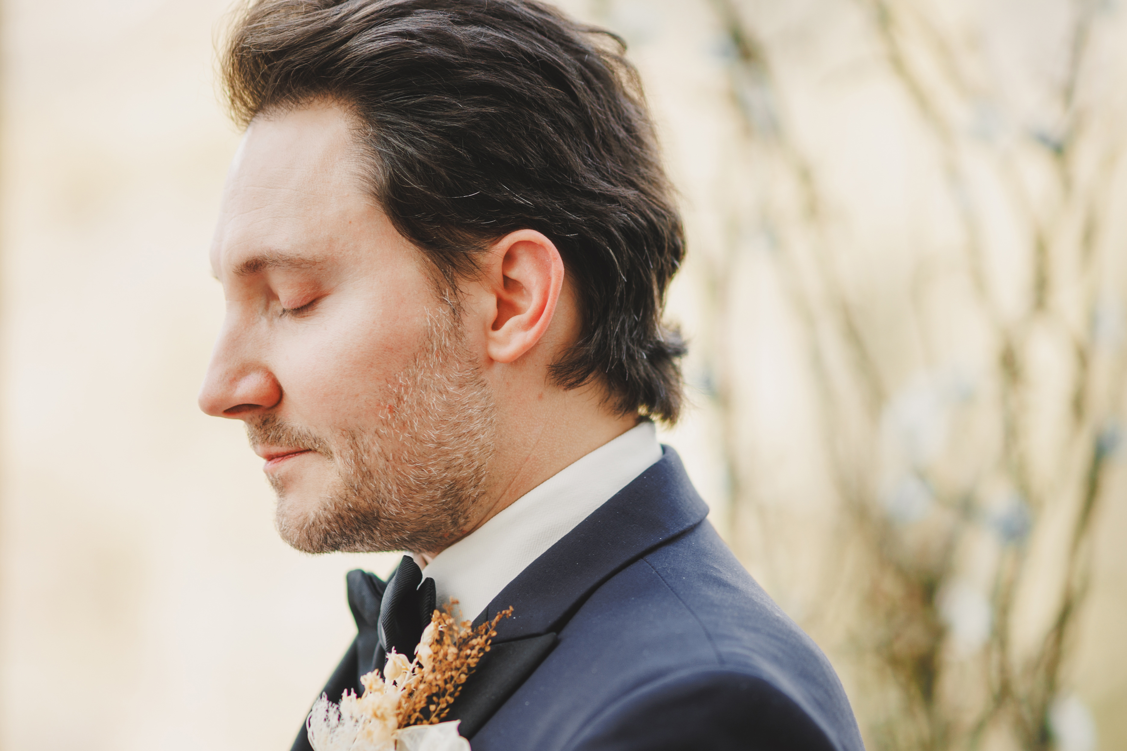 A man in a suit with a buttonhole stands with eyes closed, facing left. The background is softly blurred with light colours.