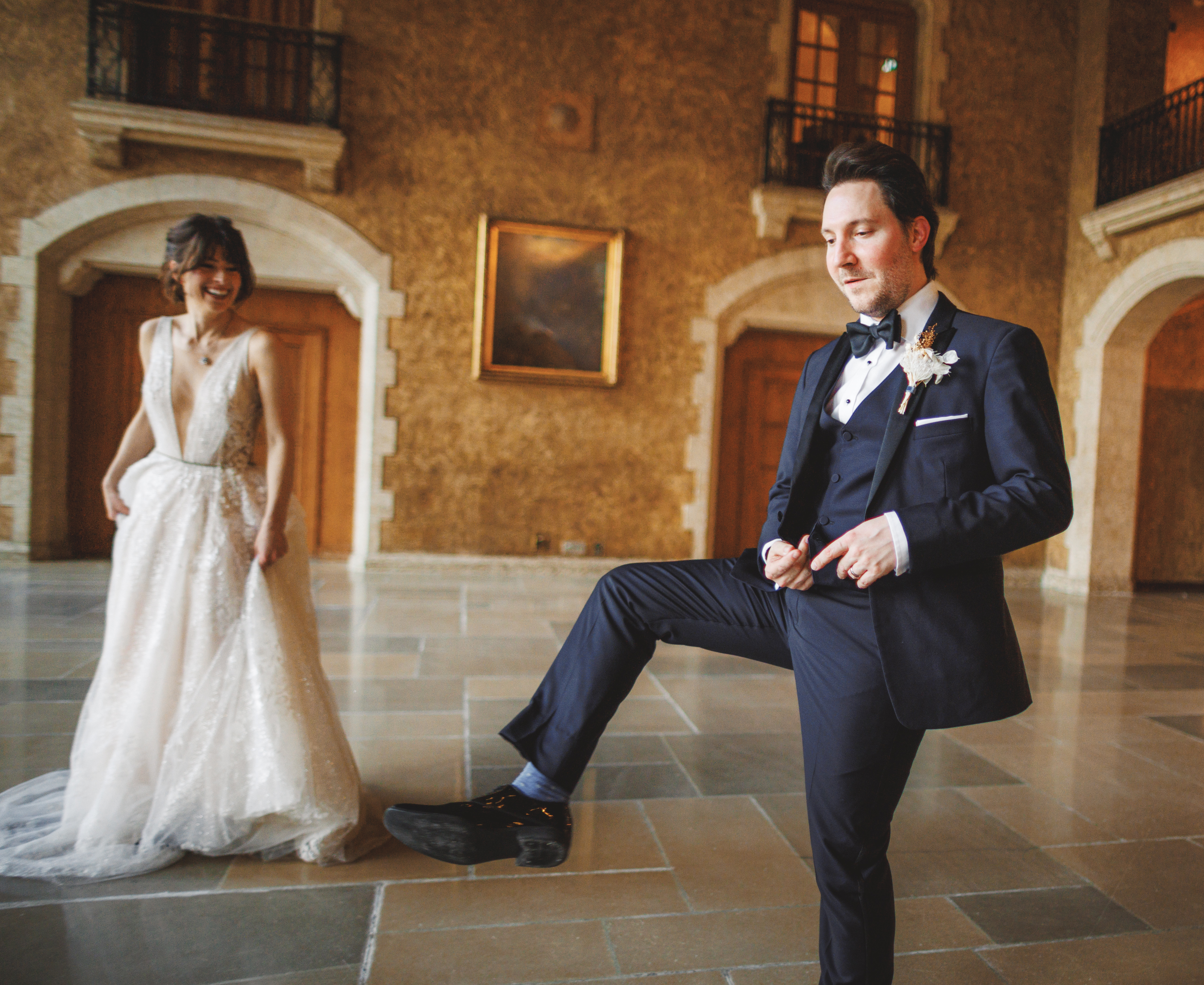 A bride in a white dress smiles whilst a groom in a dinner suit poses, lifting one foot, inside a spacious hall with stone walls and wooden doors.