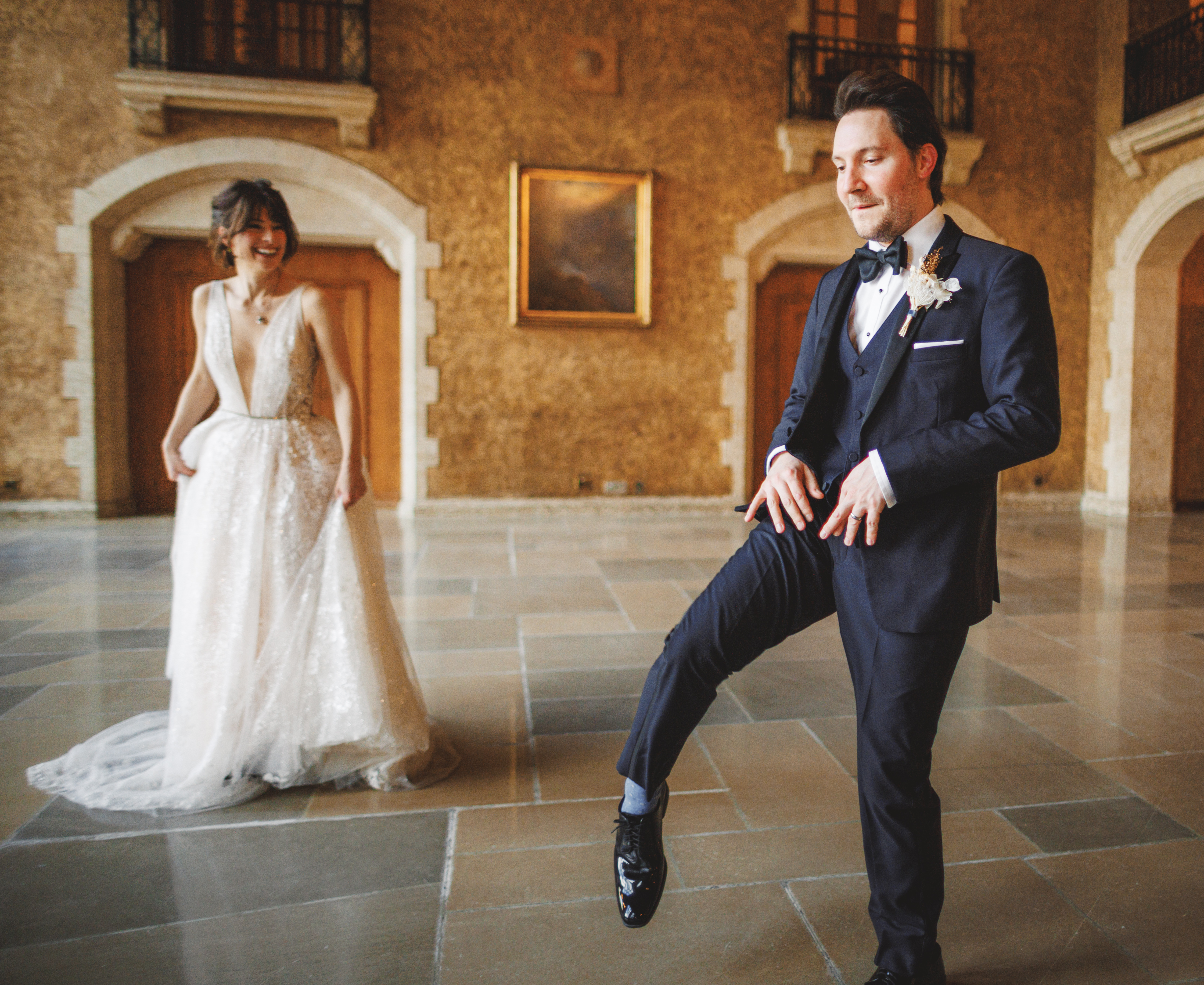 A bride in a white dress smiles as a groom in a black dinner suit playfully lifts his leg in a large, elegant hall with stone floors and wooden doors.