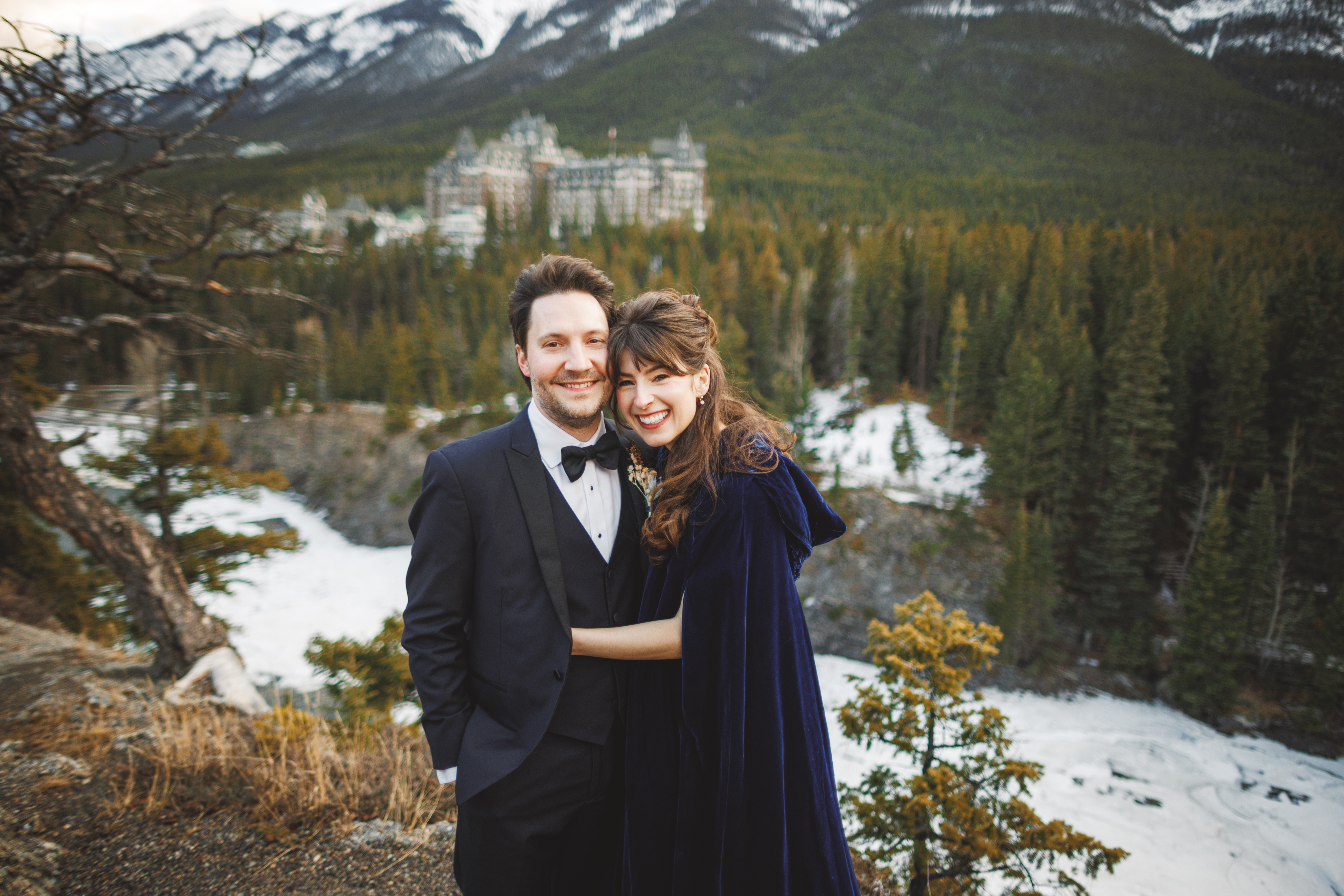 A smiling couple in formal attire pose outdoors with snow, trees, and mountains in the background. A large building is visible among the trees.