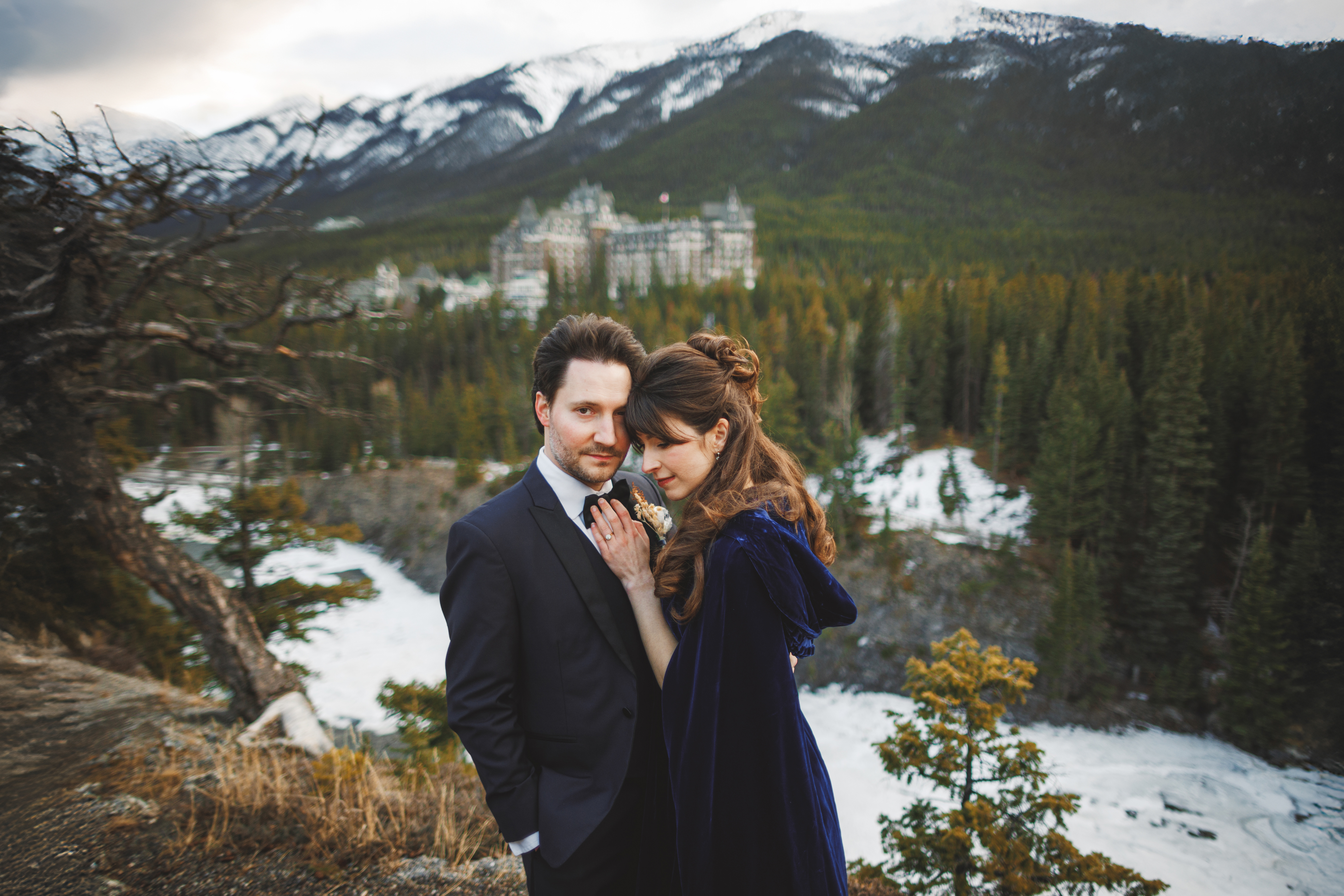 A man in a suit and a woman in a dark cloak stand close together outdoors with a forest, a river, and mountains in the background.