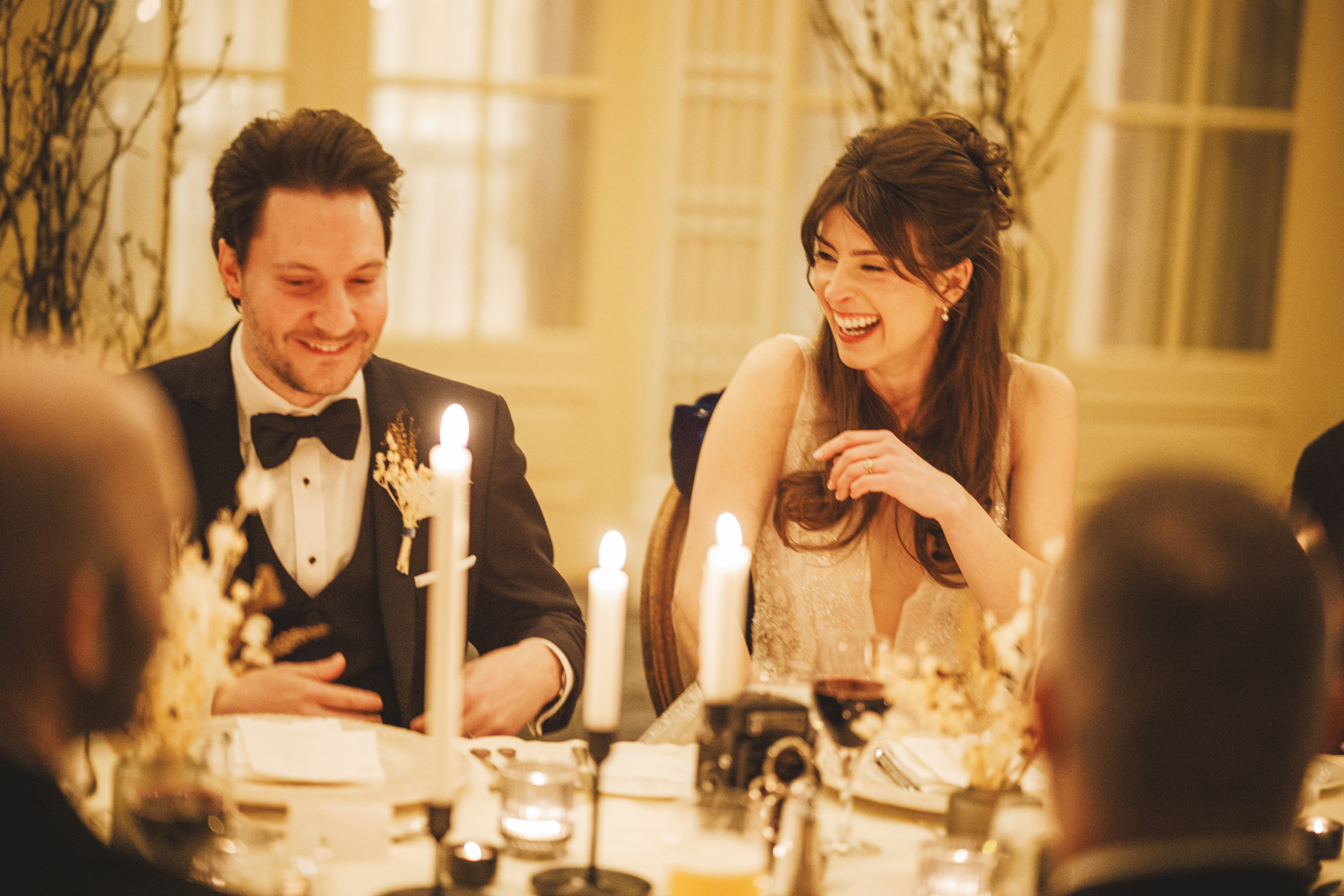 A man in a dinner jacket and a woman in a formal dress sit at a candlelit table, smiling and laughing during a formal event.