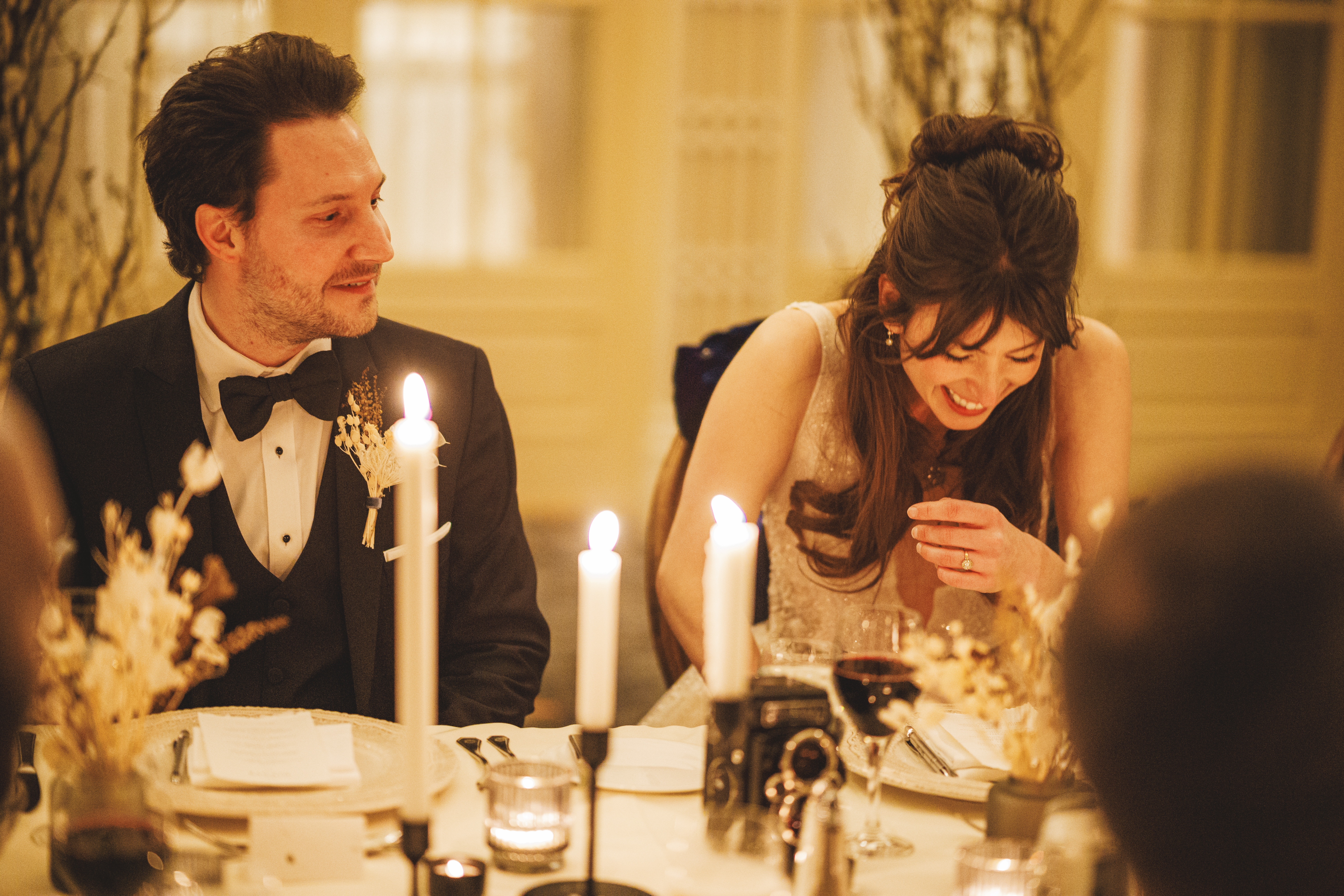 A man in a suit and a woman in a white dress sit at a decorated table with candles, laughing and sharing a moment during a formal event.