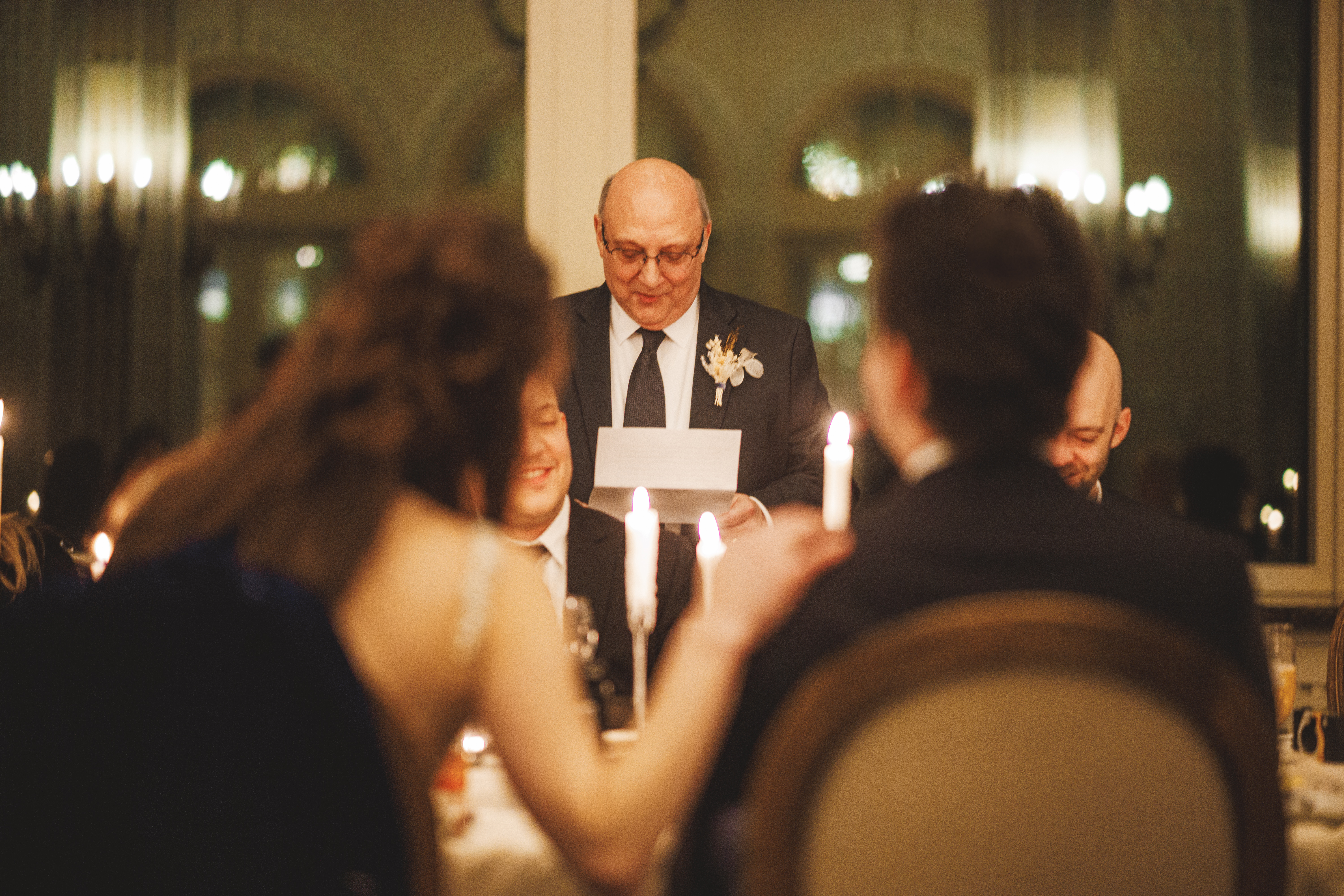A man in a suit stands and reads from a sheet of paper at a formal dinner, with guests seated round a table with lit candles.