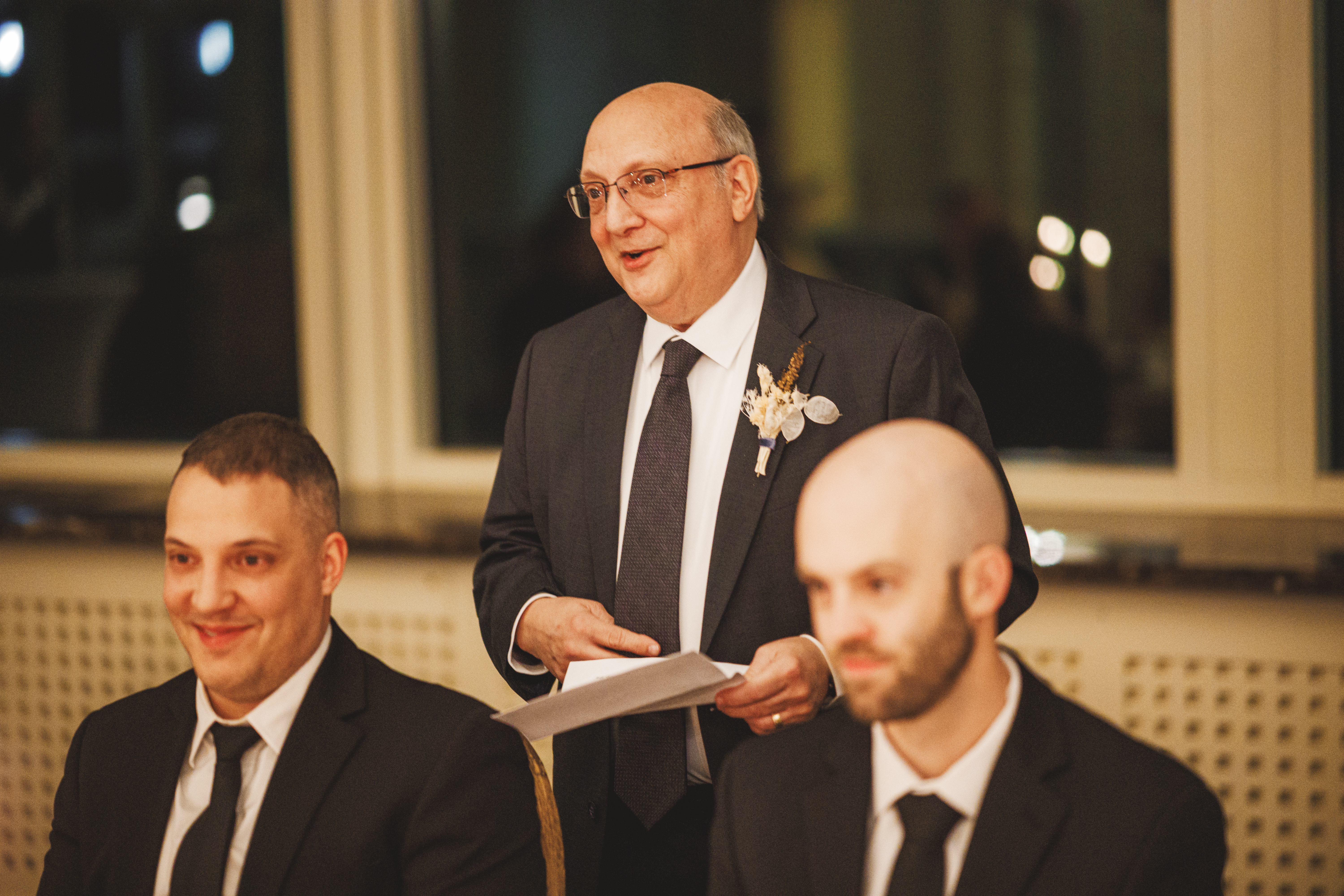 An older man in a suit delivers a speech while holding papers, standing behind two seated men in suits at a formal indoor event.