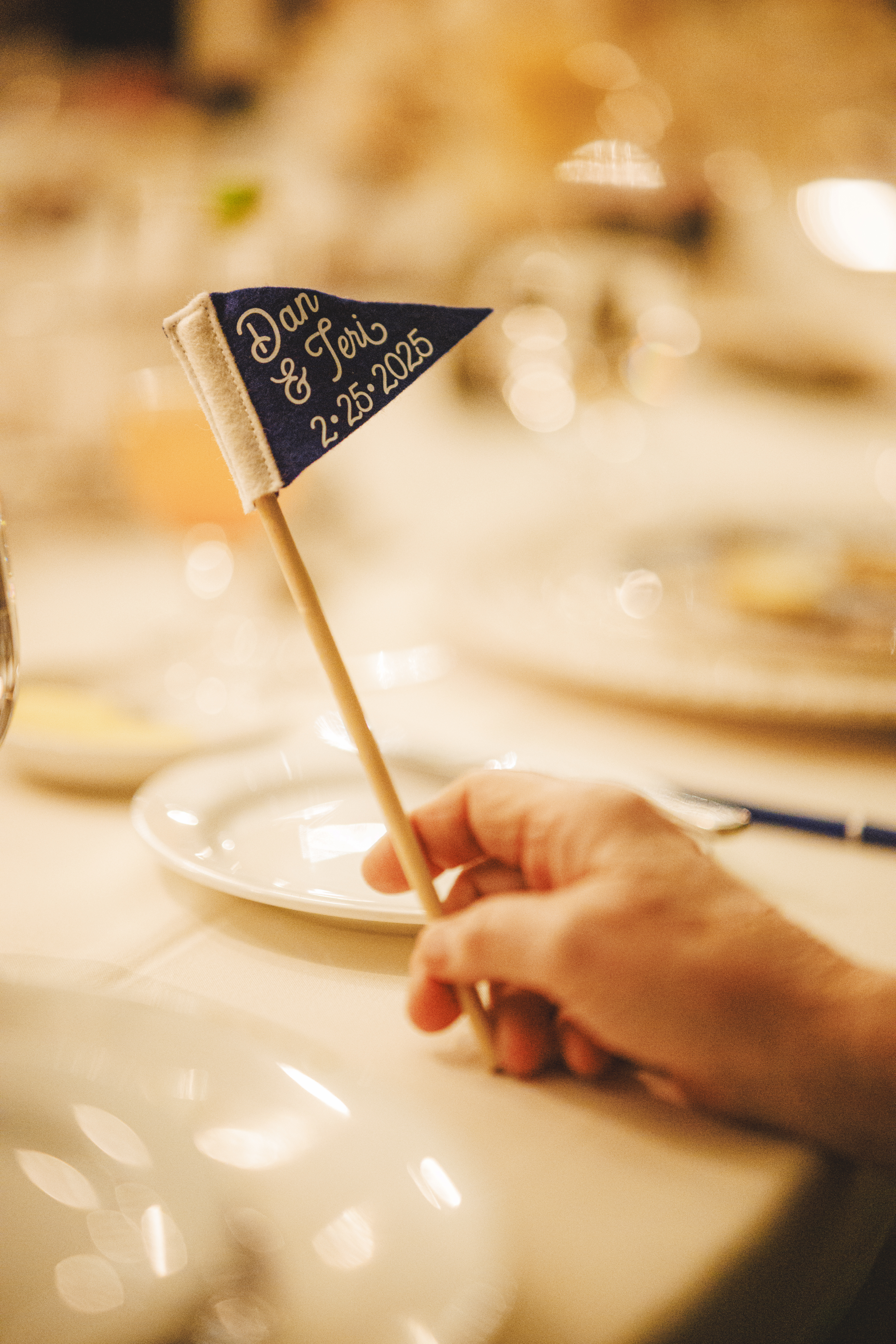 A hand holds a small blue pennant flag reading “Dan & Jeri’s 2.25.2025” at a set dining table, capturing the intimate charm of their Banff Wedding at the iconic Fairmont Hotel Banff.