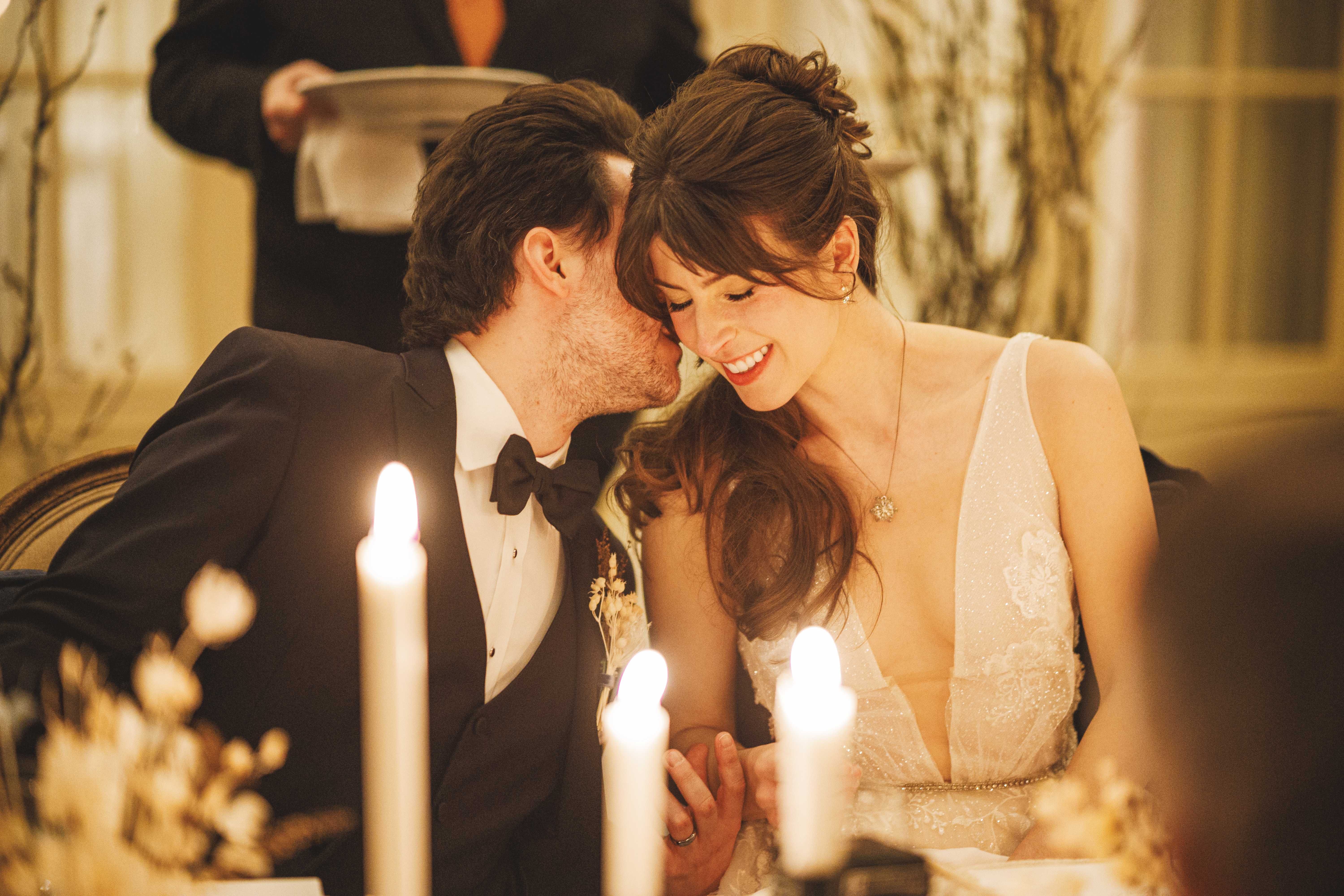 A groom in a black tuxedo kisses the bride on the cheek as she smiles, seated at a candlelit table during a wedding reception.