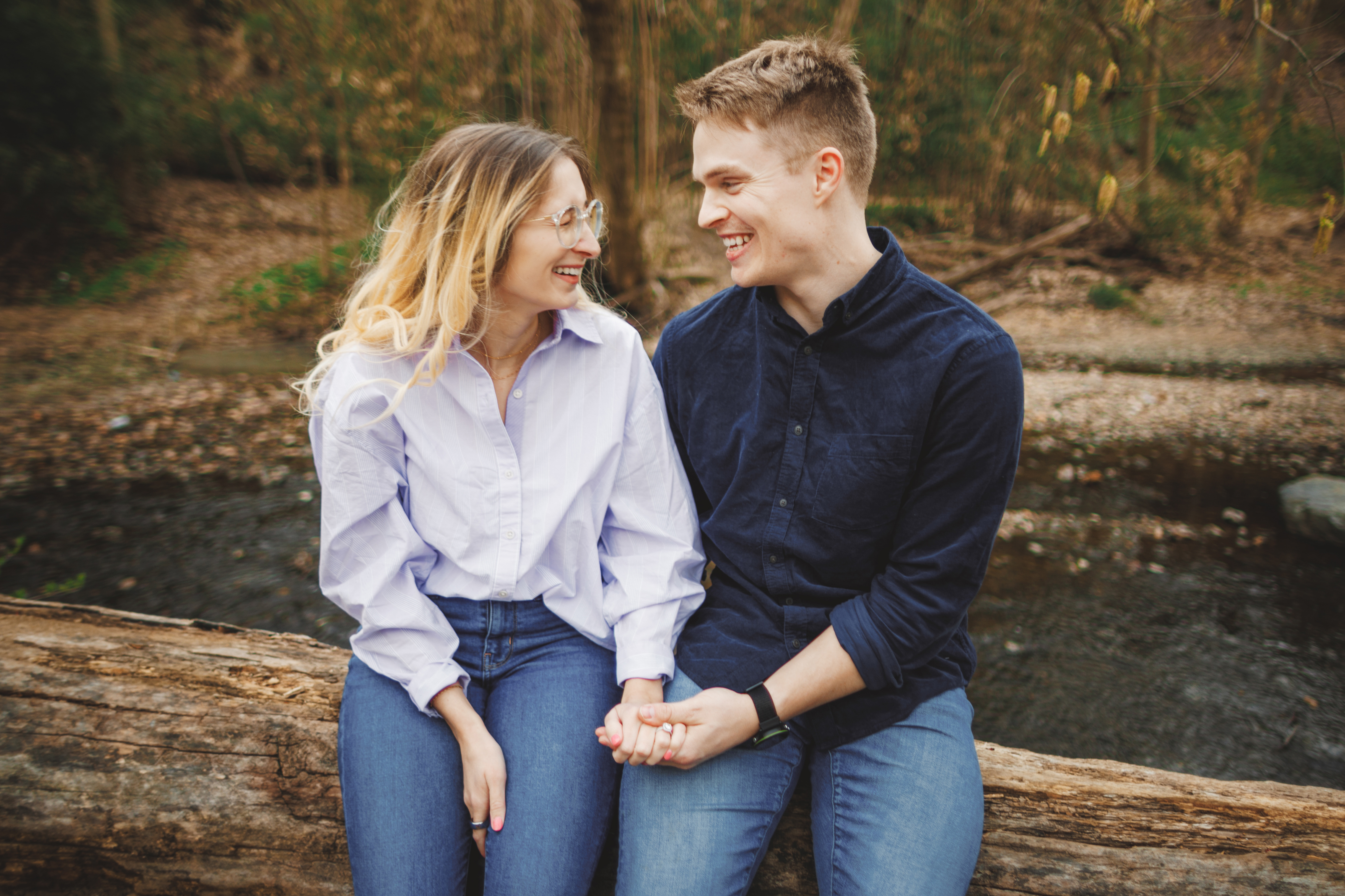 A man and woman sit on a log outdoors, holding hands and smiling at each other, with trees and a stream in the background.