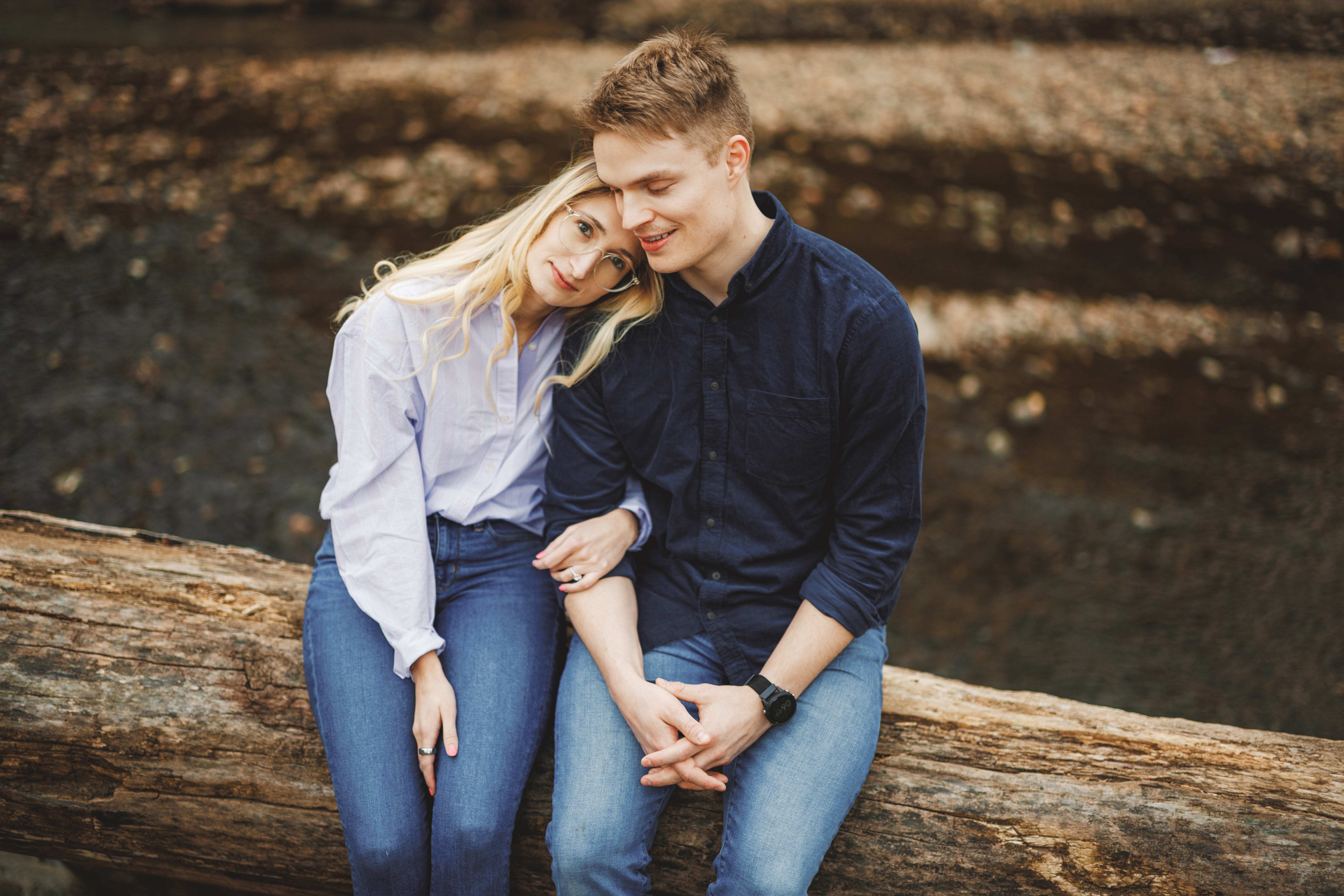 A woman rests her head on a man's shoulder whilst they sit on a log outdoors, holding hands and wearing casual clothes.