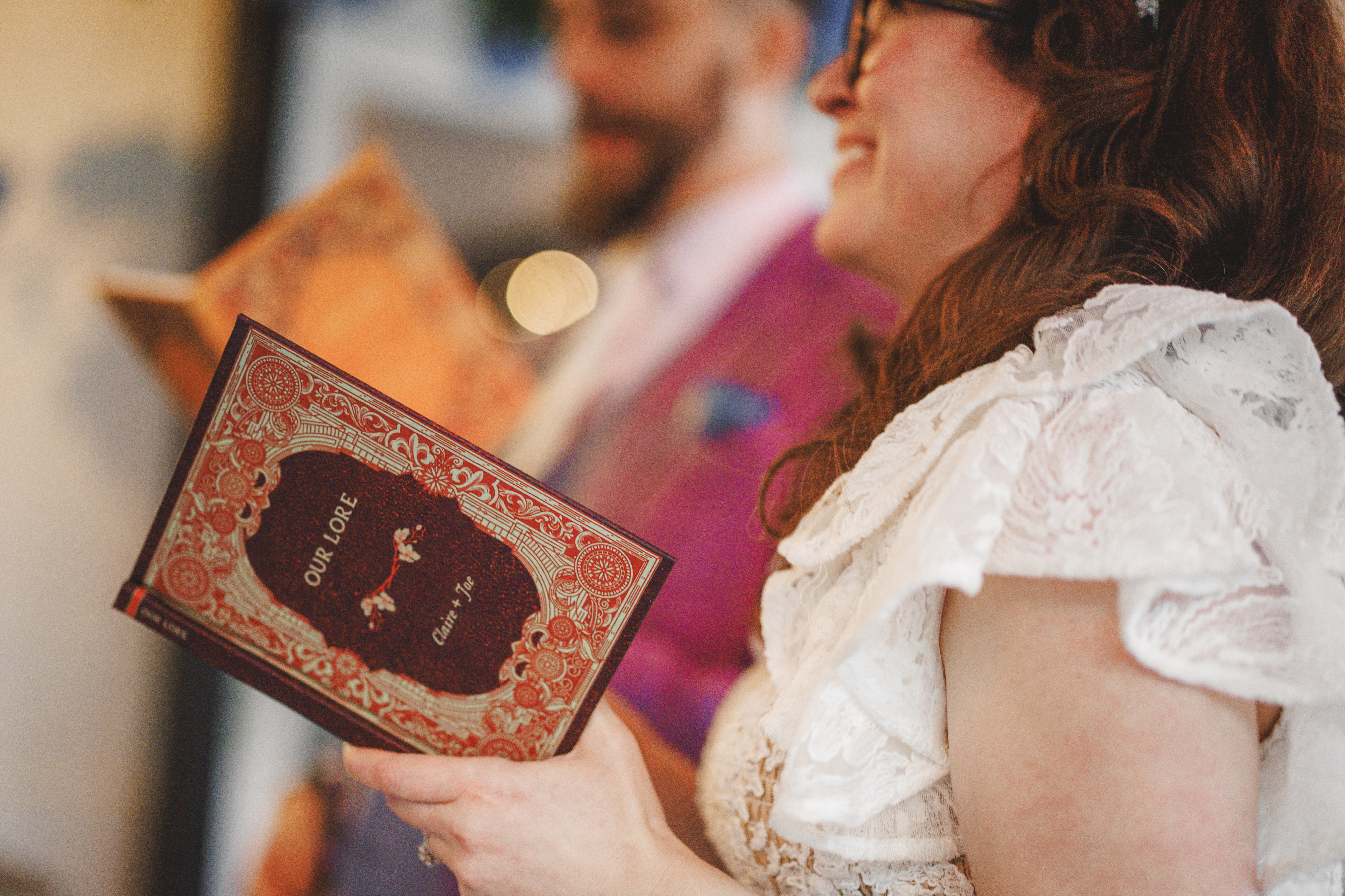 A person in a white lace dress holds a red, ornate book titled "Our Love" whilst standing beside another person in a purple suit.