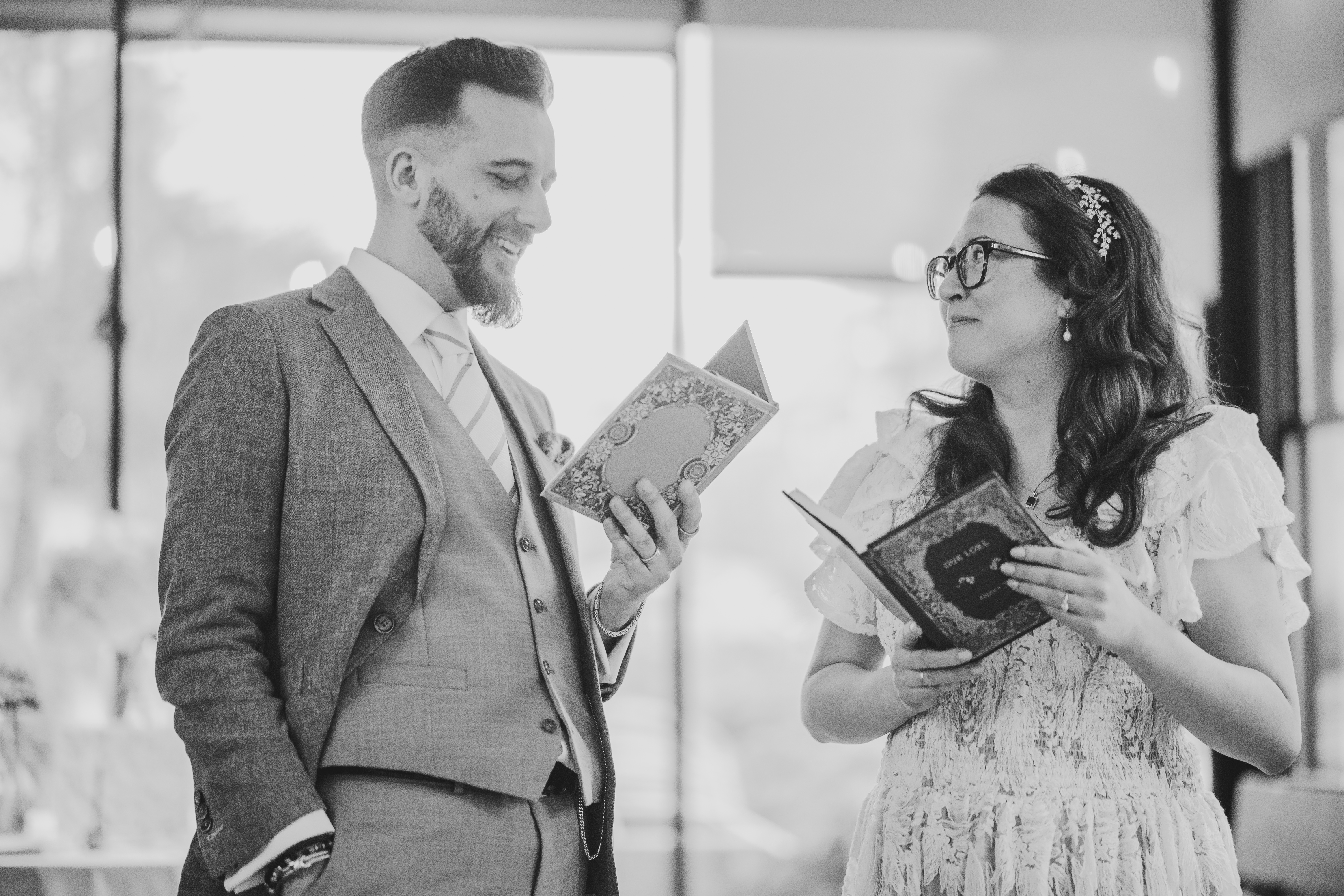 A man in a suit and a woman in a white dress stand facing each other, each holding an open book and smiling, in a well-lit indoor setting.