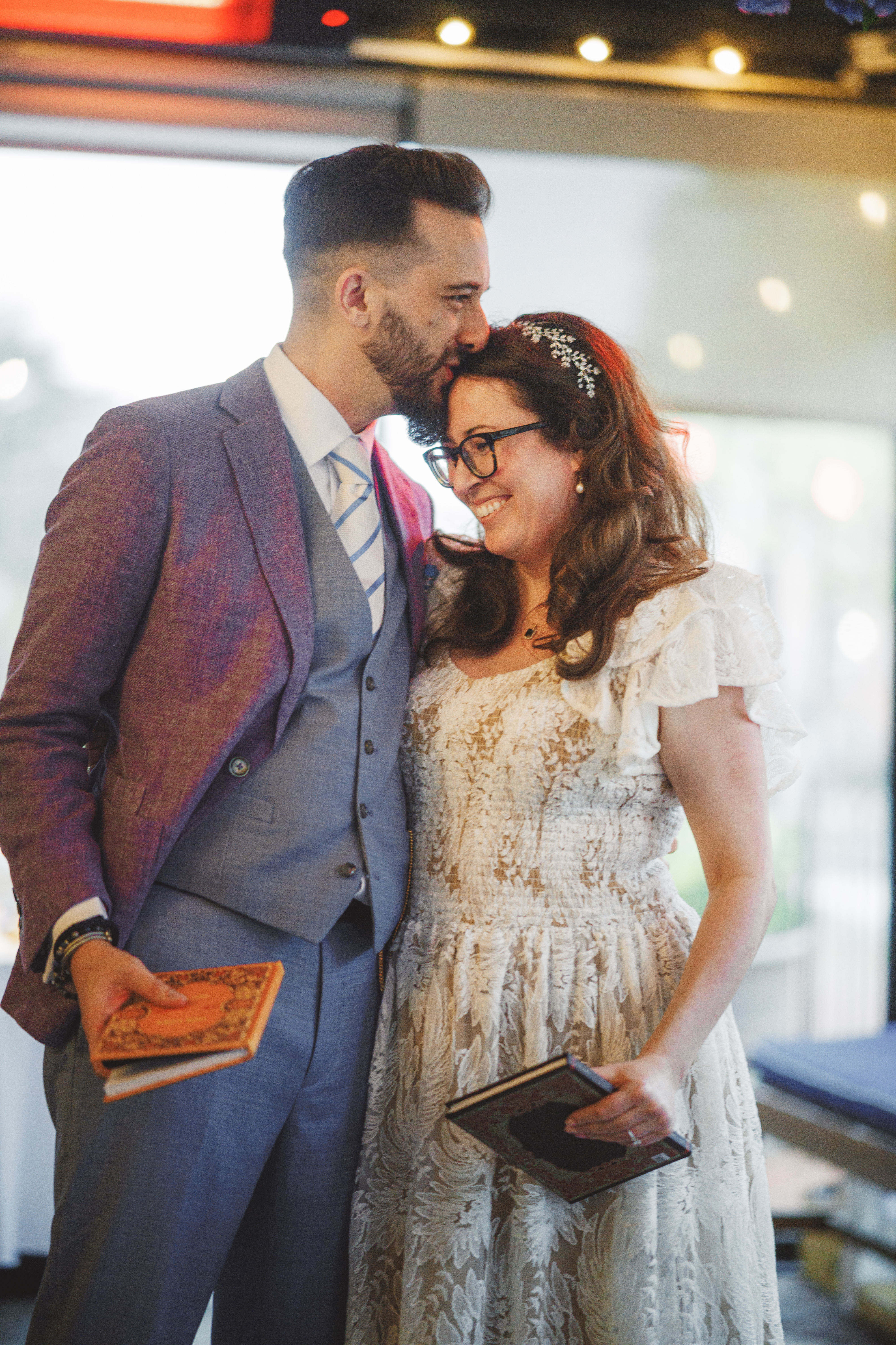 A man in a suit kisses a smiling woman in a white dress on the head. Both are holding books and standing close together indoors.