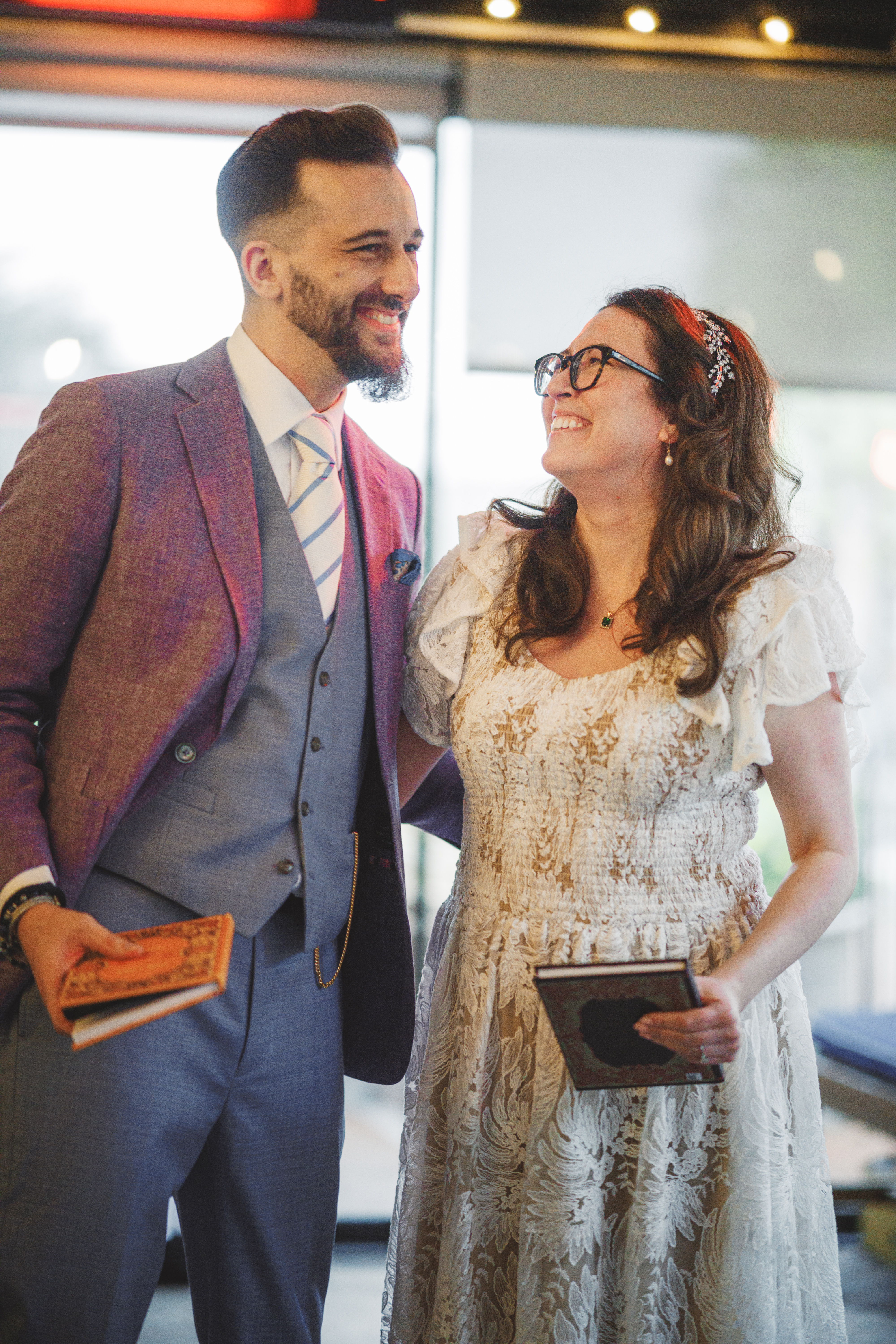 A man in a suit and a woman in a lace dress smile at each other whilst holding books indoors.