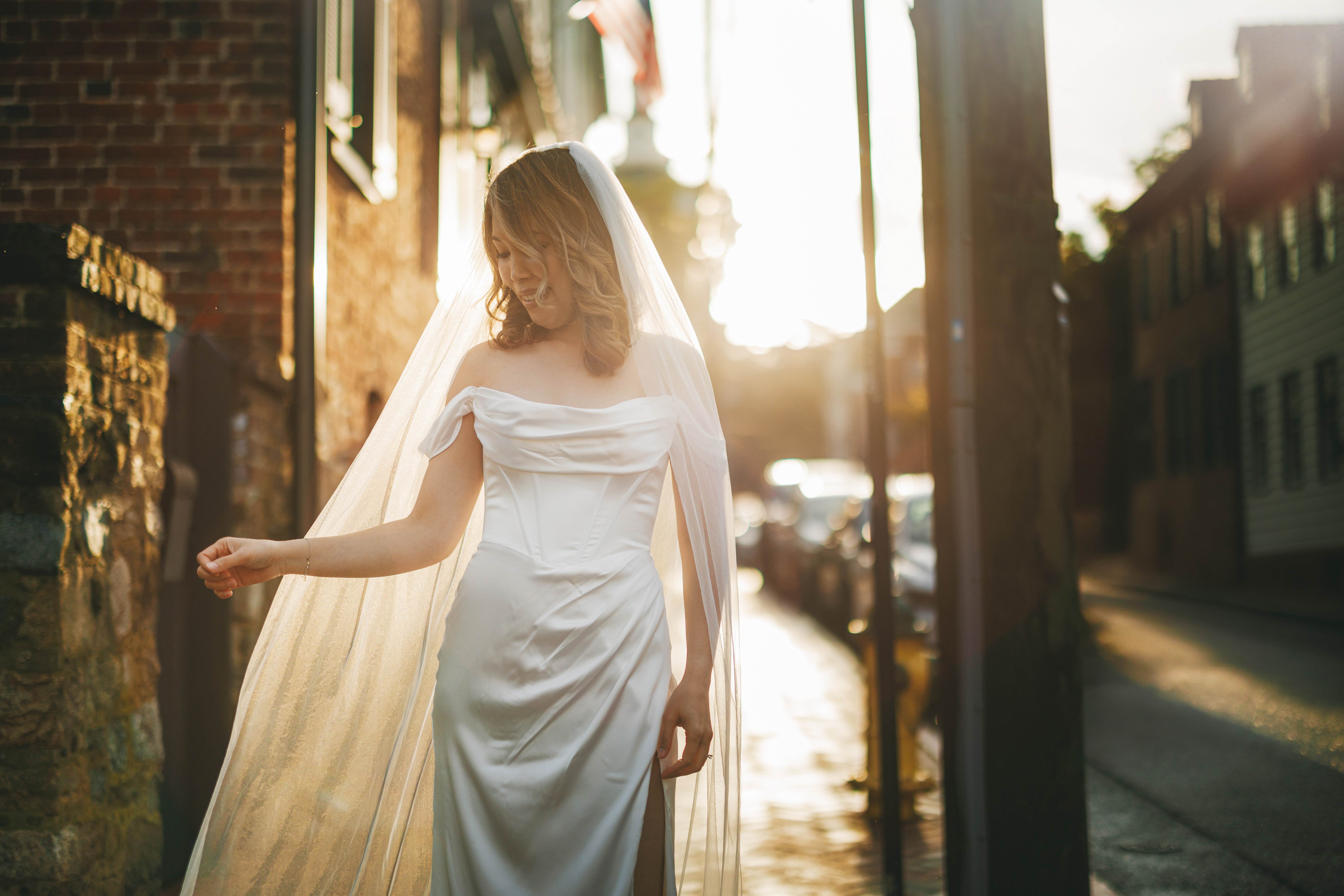A bride in an off-the-shoulder white gown and veil stands on a sunlit street, touching her veil, with buildings and parked cars in the background.