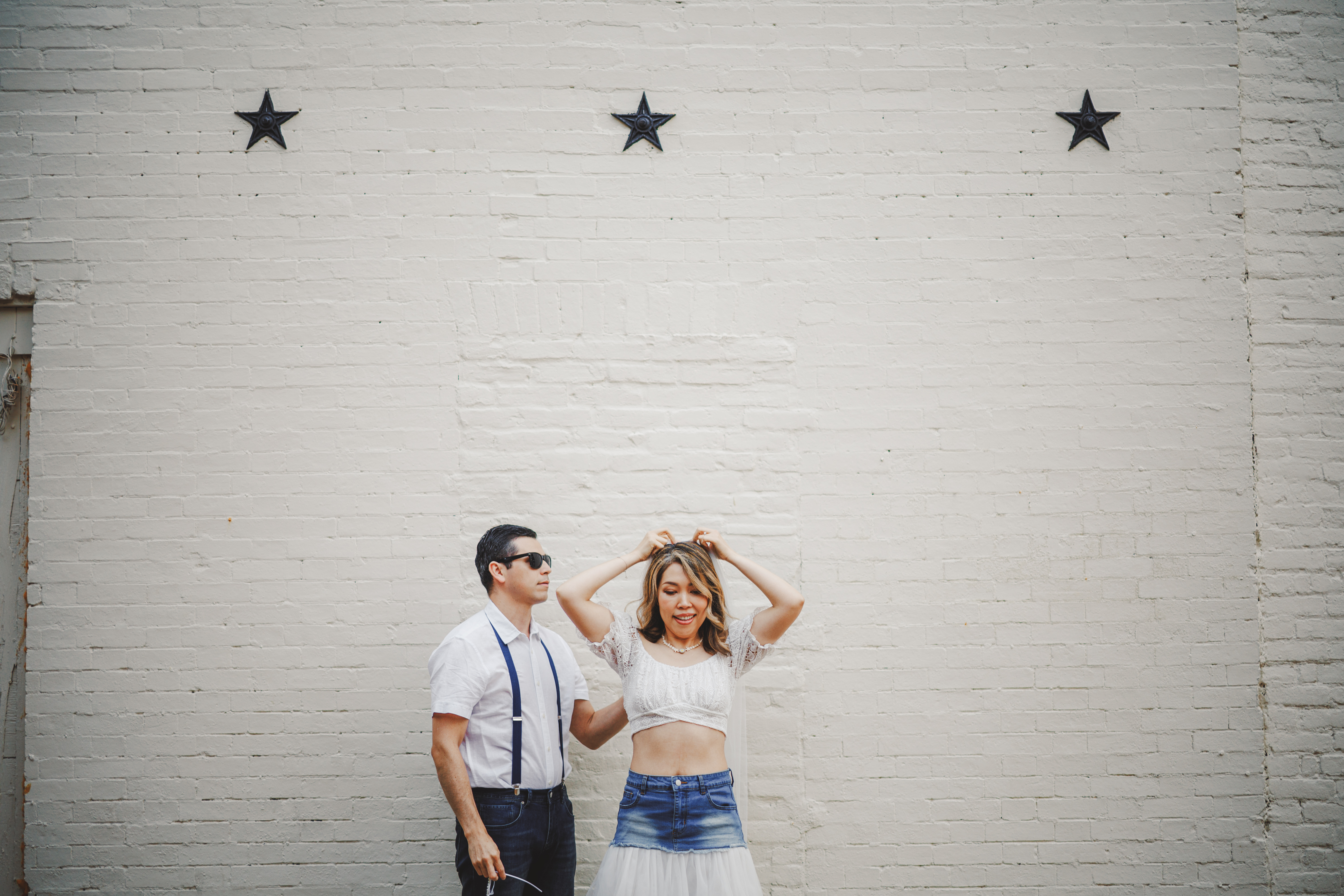 A man and woman stand in front of a white brick wall with three black stars above them; the man is looking forward whilst the woman poses with her hands on her head.