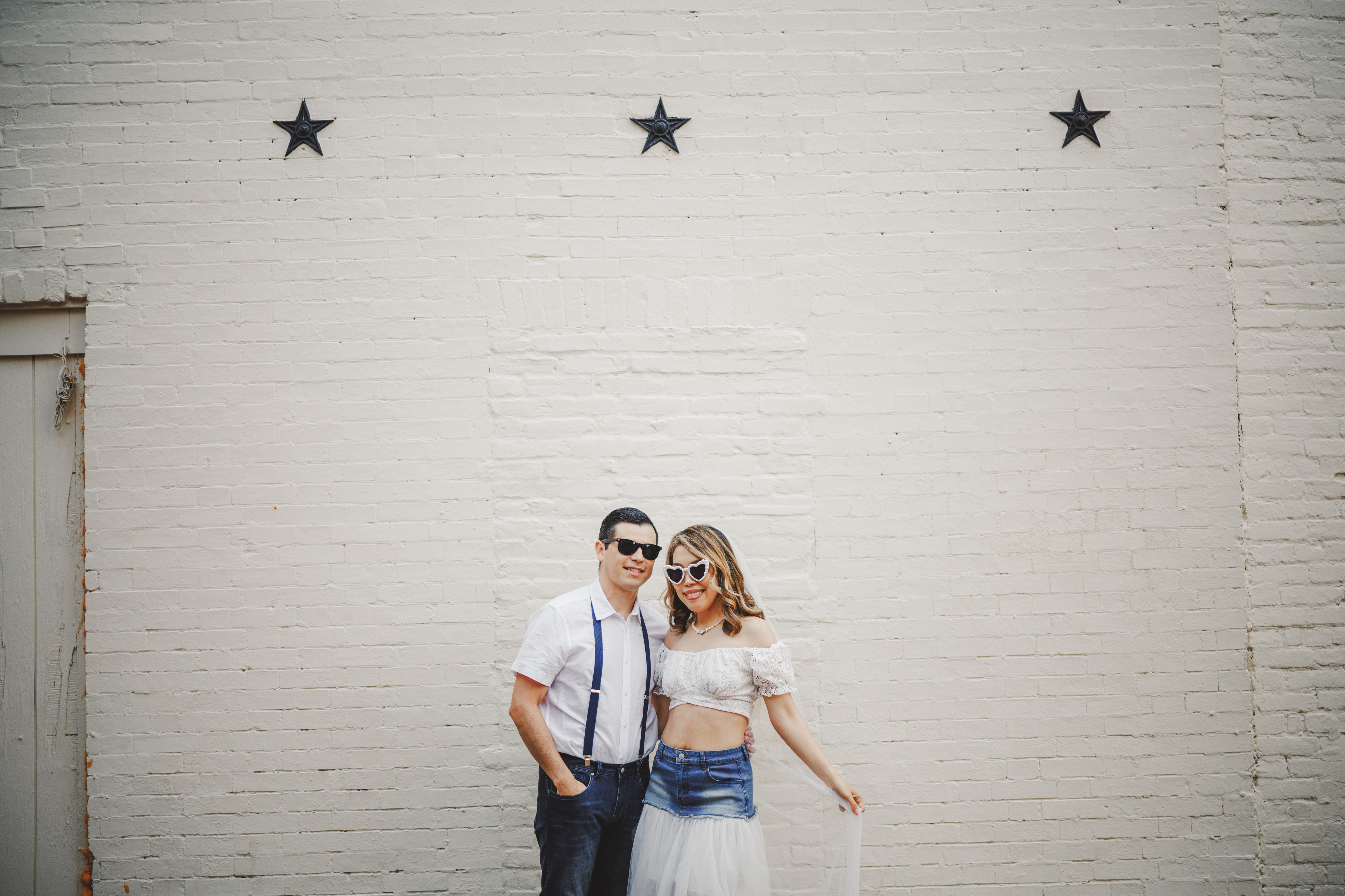 A man and woman wearing white tops and sunglasses stand smiling together in front of a white brick wall with three black stars above them.