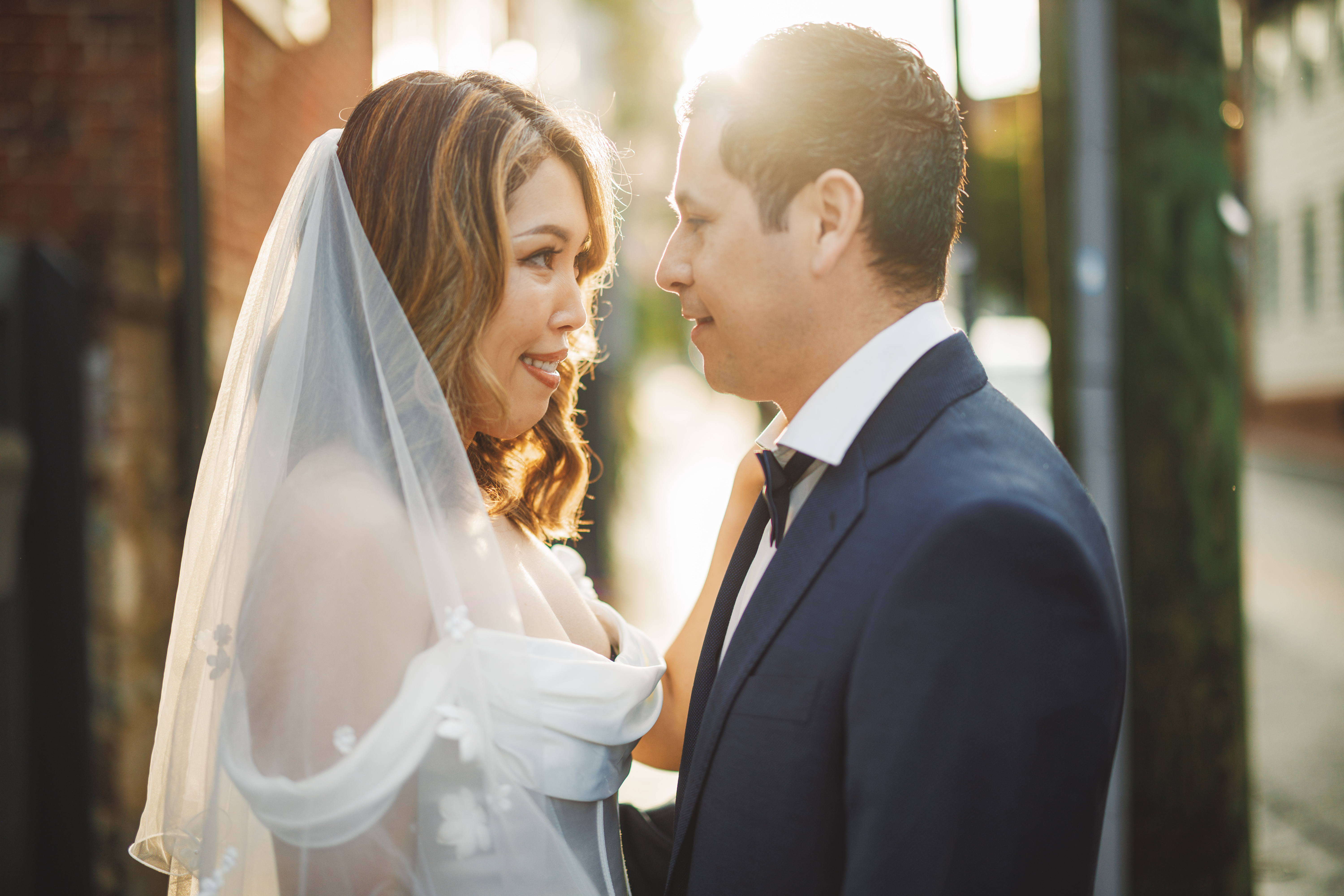 A bride and groom face each other outdoors in formal attire, with sunlight shining between them.