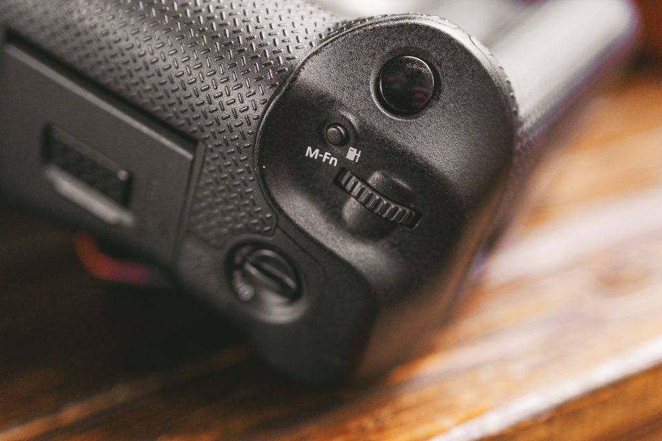 Close-up of a textured camera grip showing a control dial, M-Fn button, and part of a switch, resting on a wooden surface. Close-up of a textured camera grip showing a control dial, M-Fn button, and part of a switch, resting on a wooden surface.