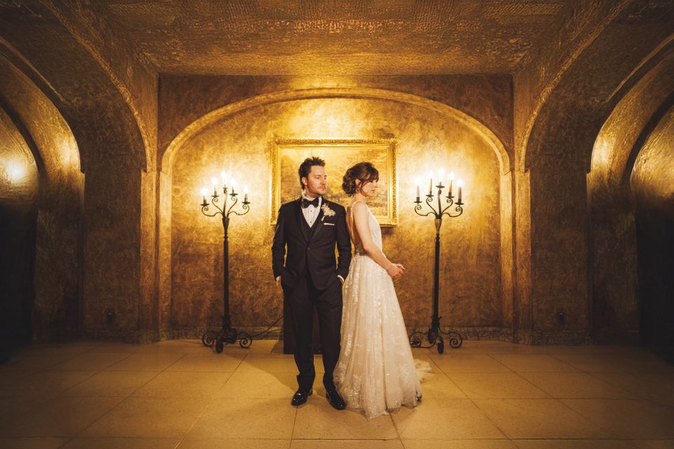 A couple in formal wedding attire stands back-to-back in an ornate room with gold walls, arched ceilings, candelabras, and a framed painting in the background. A couple in formal wedding attire stands back-to-back in an ornate room with gold walls, arched ceilings, candelabras, and a framed painting in the background.