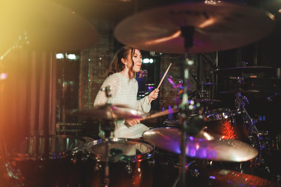 A woman in a light sweater plays a drum set on stage, holding drumsticks and smiling, with stage lights and drum equipment visible around her. A woman in a light sweater plays a drum set on stage, holding drumsticks and smiling, with stage lights and drum equipment visible around her.