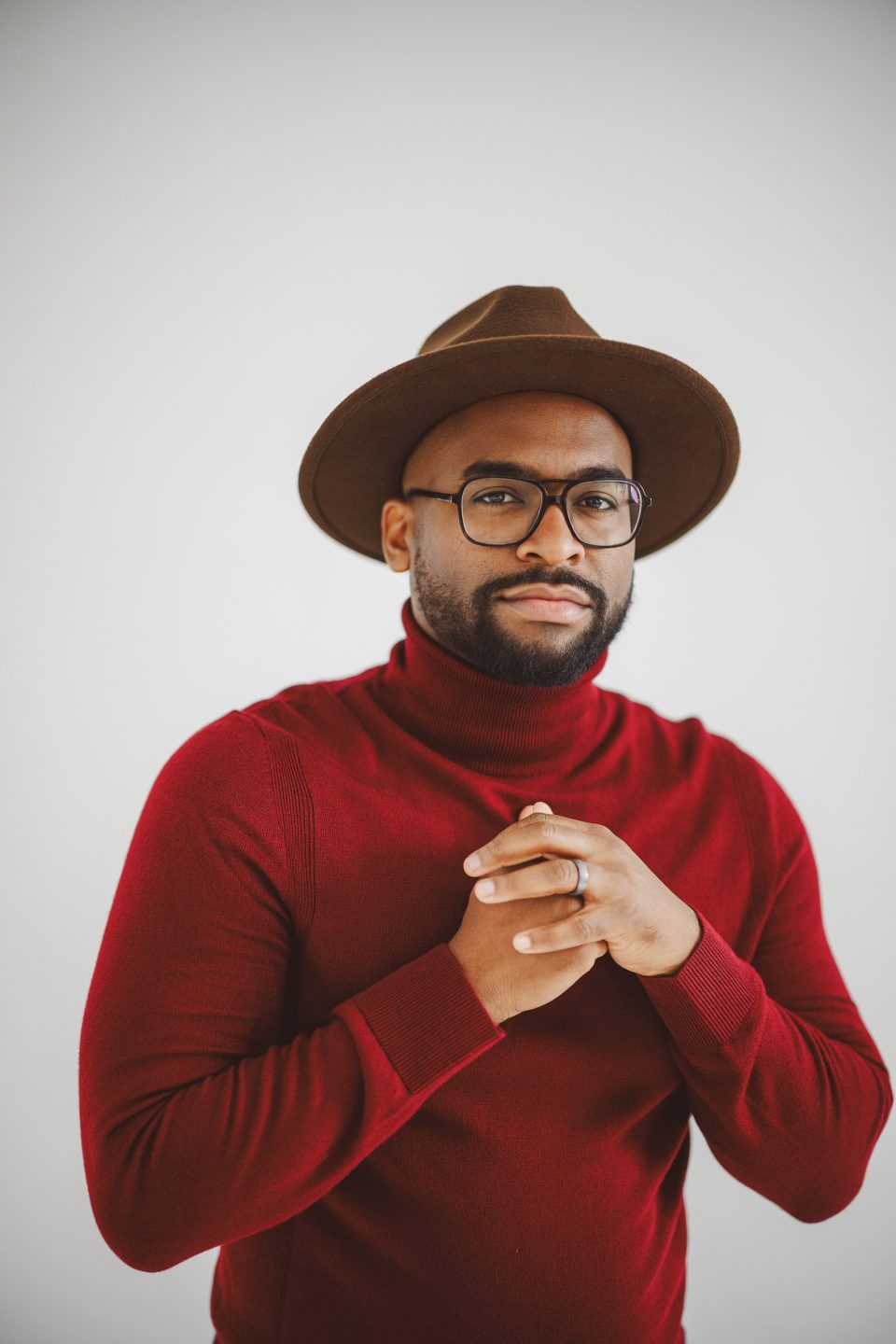 A man wearing glasses, a brown hat, and a red turtleneck sweater stands against a plain light background with his hands clasped together. A man wearing glasses, a brown hat, and a red turtleneck sweater stands against a plain light background with his hands clasped together.