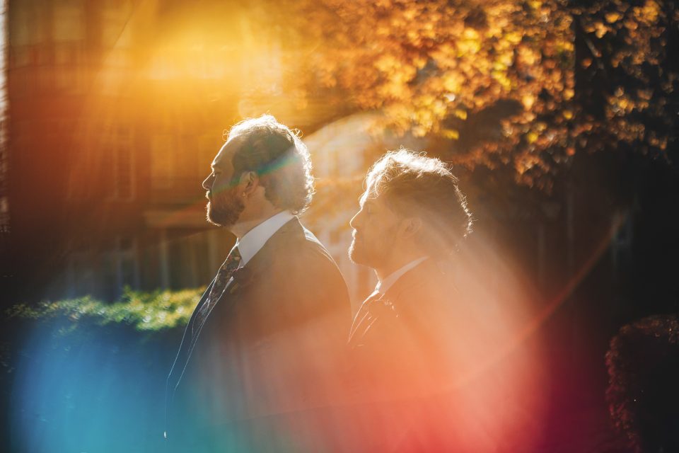 Two men in suits stand outdoors in sunlight, surrounded by colorful lens flares and autumn foliage in the background. Two men in suits stand outdoors in sunlight, surrounded by colorful lens flares and autumn foliage in the background.