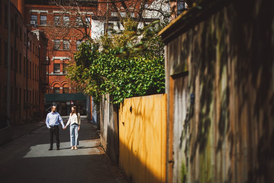 Two people walk hand in hand down a narrow urban alleyway lined with buildings and wooden fences, with sunlight casting shadows. Two people walk hand in hand down a narrow urban alleyway lined with buildings and wooden fences, with sunlight casting shadows.