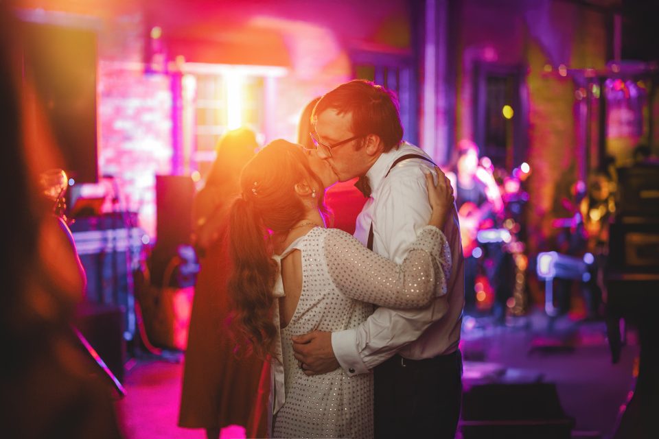 A couple dressed in formal attire share a kiss while dancing at a party or wedding reception with colorful lighting and a live band in the background. A couple dressed in formal attire share a kiss while dancing at a party or wedding reception with colorful lighting and a live band in the background.