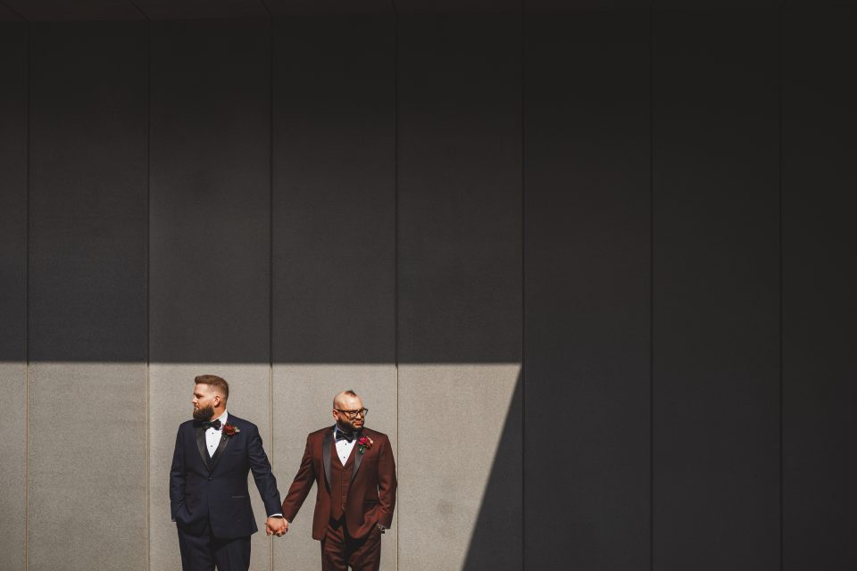 Two men in suits, one navy and one brown, stand holding hands against a tall, gray, shadowed wall with a beam of sunlight partially illuminating them. Two men in suits, one navy and one brown, stand holding hands against a tall, gray, shadowed wall with a beam of sunlight partially illuminating them.