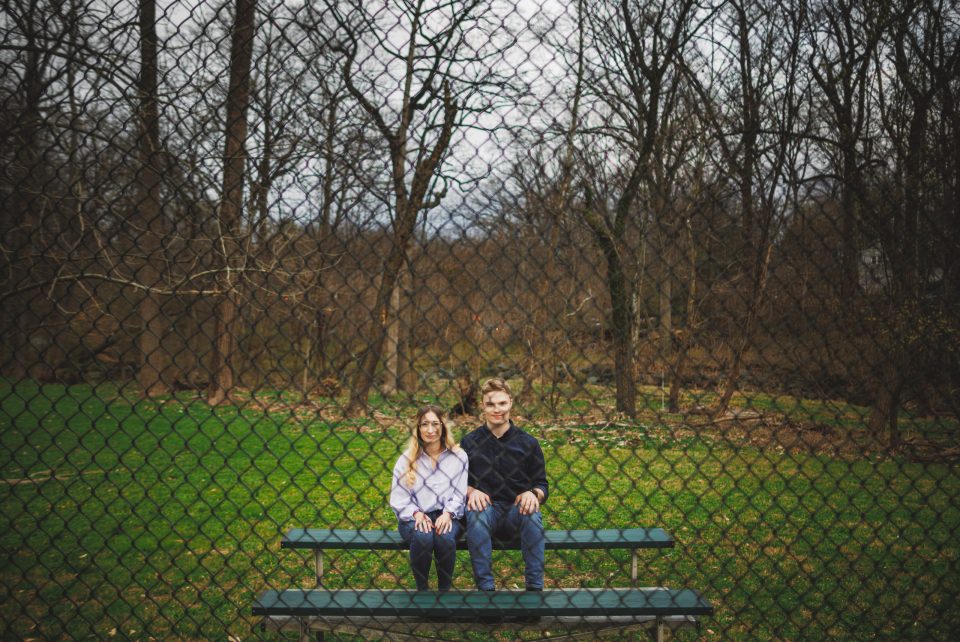 Two people sit side by side on a bench in a park, behind a chain-link fence with leafless trees and green grass in the background. Two people sit side by side on a bench in a park, behind a chain-link fence with leafless trees and green grass in the background.