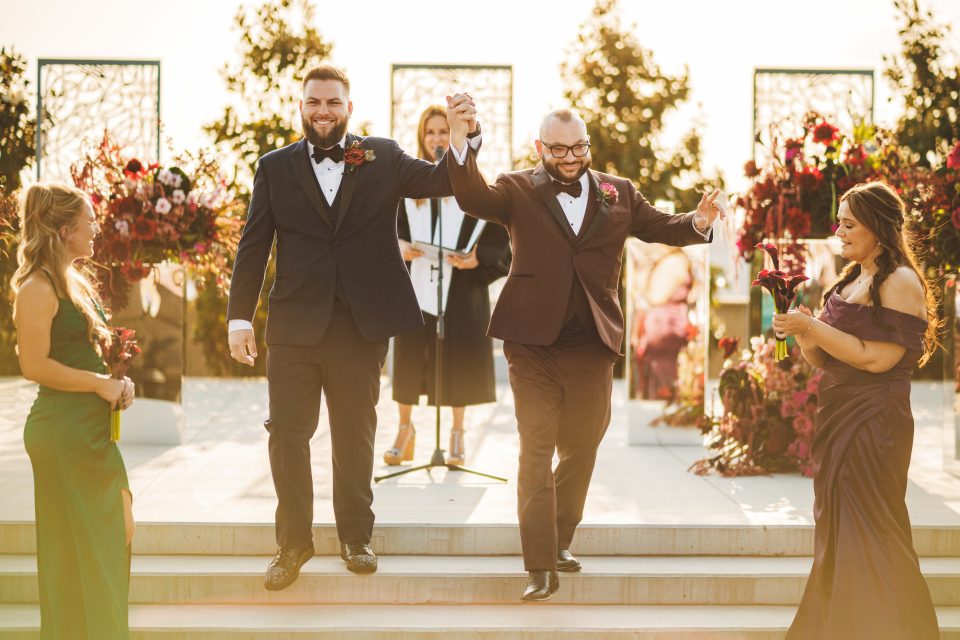 Two men in suits hold hands and smile as they walk down steps at an outdoor wedding ceremony, with two women in dresses nearby and floral arrangements in the background. Two men in suits hold hands and smile as they walk down steps at an outdoor wedding ceremony, with two women in dresses nearby and floral arrangements in the background.