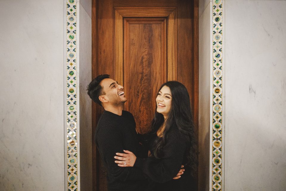 A man and a woman stand close together, smiling and laughing in front of a wooden door framed by decorative tiles. A man and a woman stand close together, smiling and laughing in front of a wooden door framed by decorative tiles.