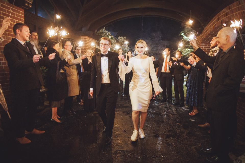 A newlywed couple walks under an archway at night as guests holding sparklers line both sides, celebrating their wedding. A newlywed couple walks under an archway at night as guests holding sparklers line both sides, celebrating their wedding.