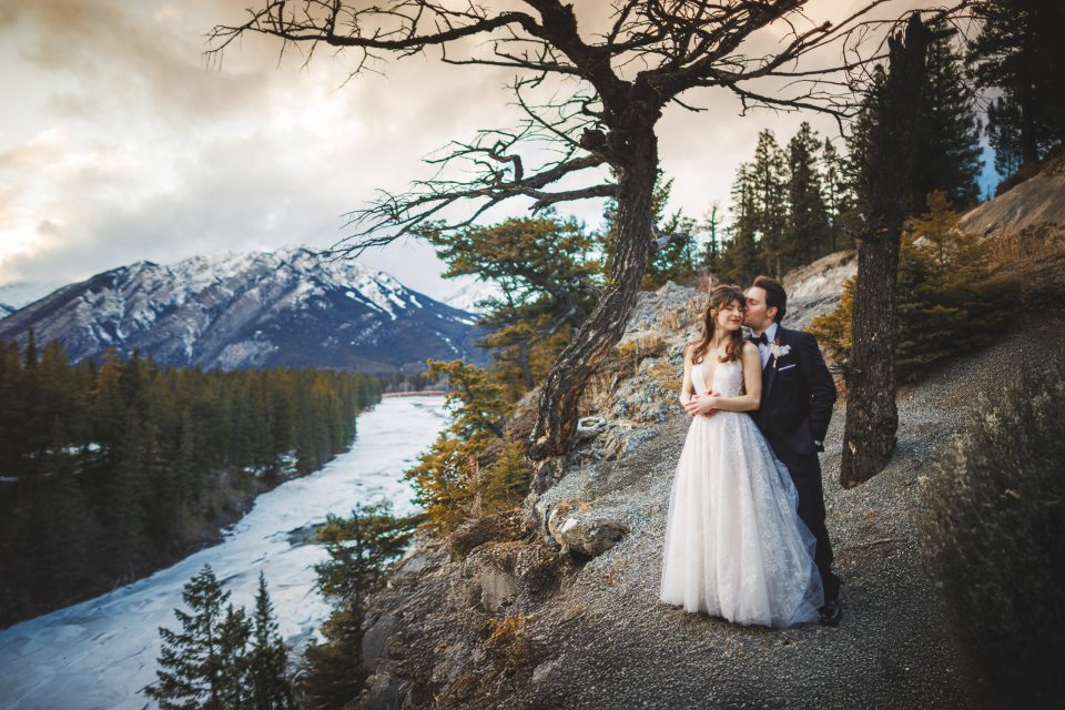 A bride and groom stand on a rocky ledge overlooking a snowy river and pine trees, with mountains in the background under a cloudy sky. A bride and groom stand on a rocky ledge overlooking a snowy river and pine trees, with mountains in the background under a cloudy sky.