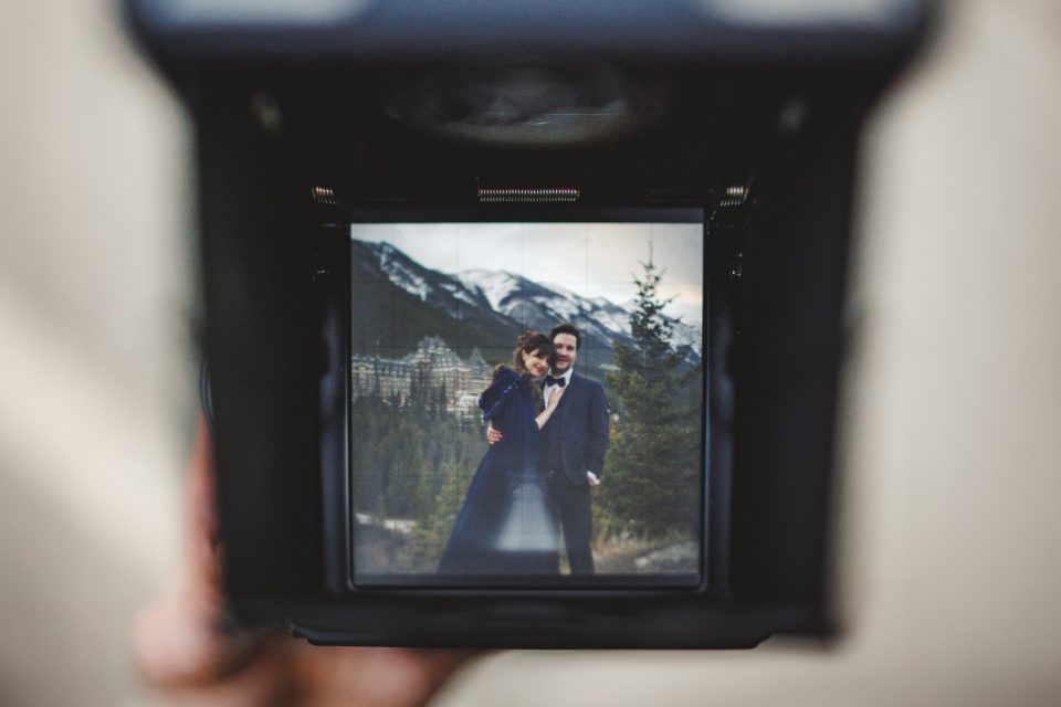 A couple poses in formal attire outdoors, with mountains and trees in the background, as seen through a camera viewfinder. A couple poses in formal attire outdoors, with mountains and trees in the background, as seen through a camera viewfinder.