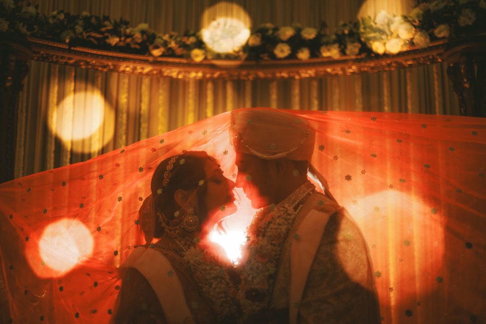 Two people in traditional wedding attire stand close under a veil, facing each other, with warm lighting and floral decorations in the background. Two people in traditional wedding attire stand close under a veil, facing each other, with warm lighting and floral decorations in the background.