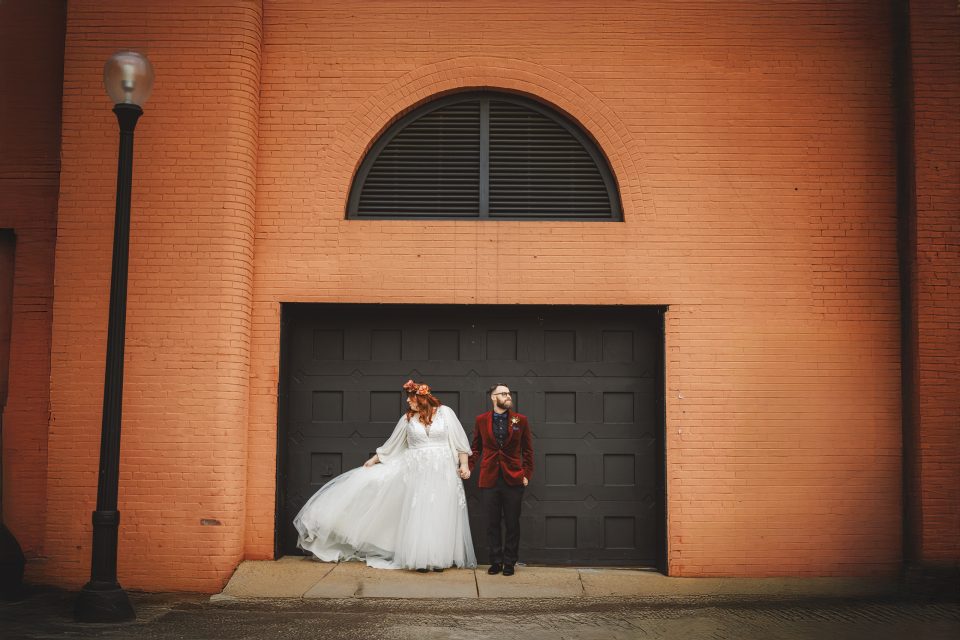 A bride in a white dress and a groom in a red suit stand side by side in front of a large black garage door set in an orange brick building. A bride in a white dress and a groom in a red suit stand side by side in front of a large black garage door set in an orange brick building.